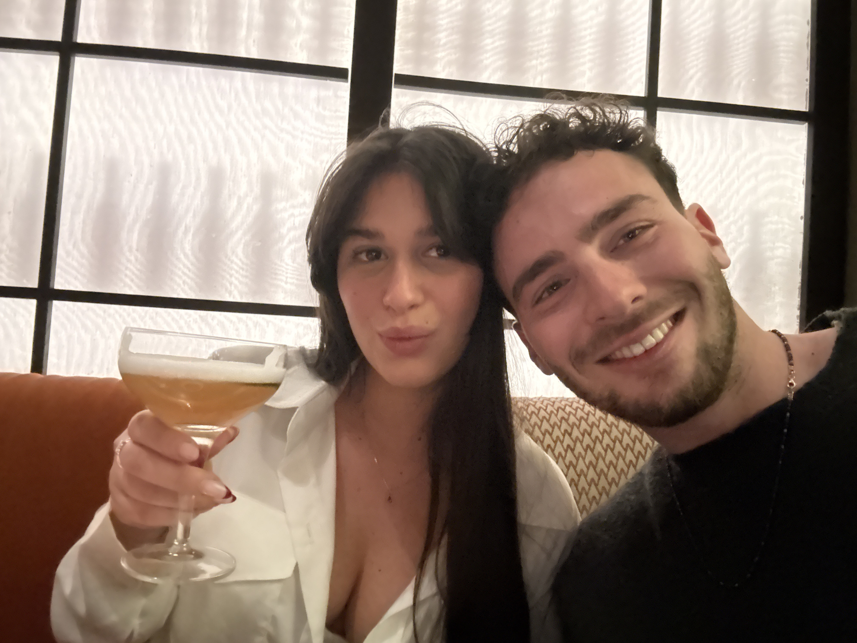 A close-up selfie captures two individuals, a woman and a man, seated indoors, likely in a bar, restaurant, or lounge setting. Both are looking directly at the camera.
The woman is on the left side of the frame, holding a cocktail glass in her right hand, raised slightly towards the camera. She has long, dark hair styled with bangs and is wearing a white collared shirt that is open at the neckline, revealing a delicate necklace with a small pendant. Her nails are manicured with dark red tips. She has dark eyes and her lips are slightly pursed, giving a subtle smile.
The man is on the right side, leaning in towards the woman. He has short, curly dark hair and a beard. He is smiling broadly, showing his white teeth, and appears happy. He is wearing a dark-colored top, possibly black or dark gray, and a dark beaded bracelet or necklace with a small metallic charm is visible around his neck.
The background features large windows with dark frames forming a grid pattern. These windows are covered with a translucent material, possibly blinds or textured glass, which diffuses light and creates a subtle wavy or rippled texture. The lighting appears soft and warm, suggesting an evening or night setting. Behind the woman, there is a solid orange or rust-colored upholstered surface, while behind the man, a beige cushion with a repeating brown geometric pattern is visible. The overall mood of the image is joyful and relaxed. No text is visible in the image.