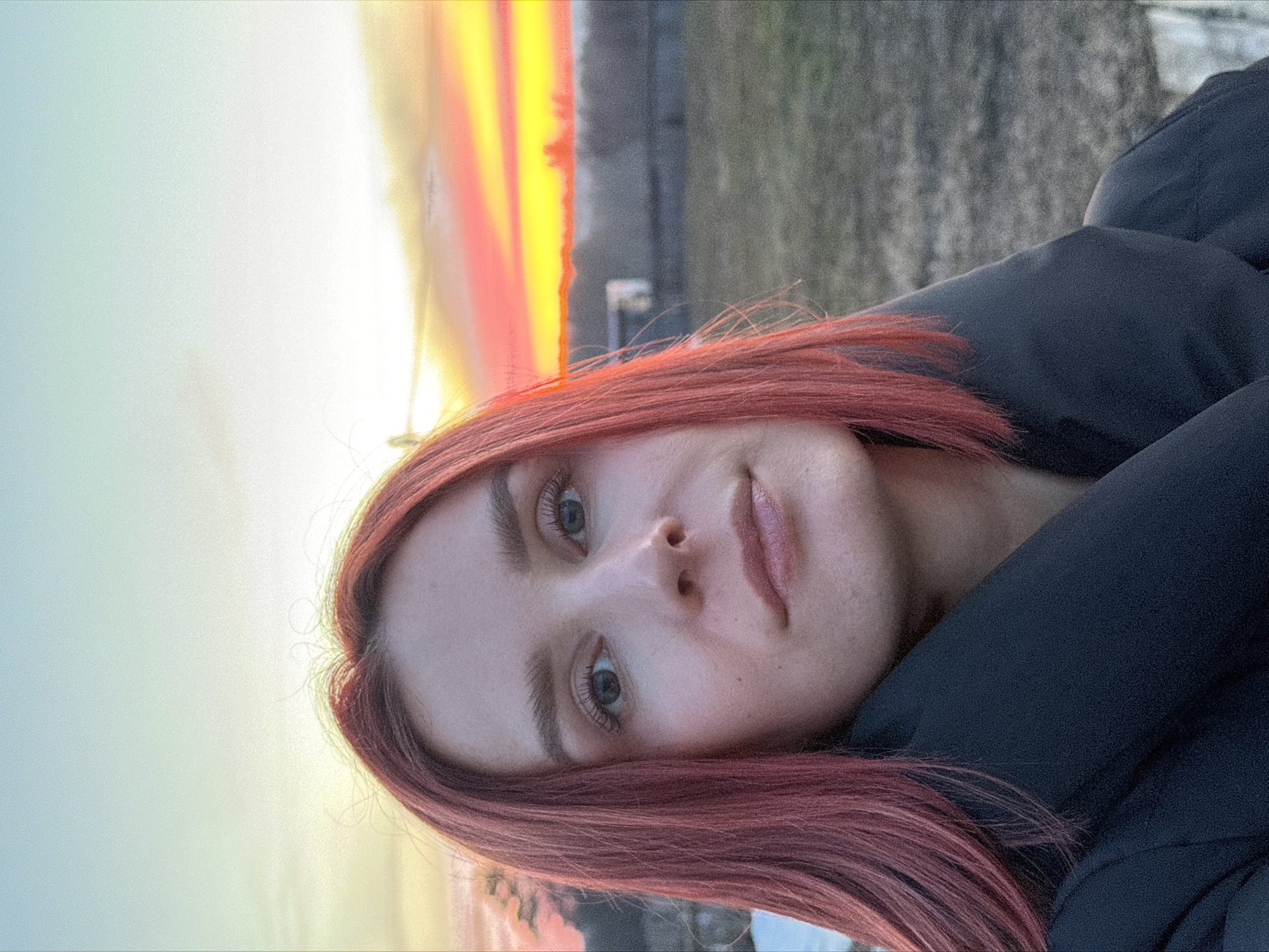 A selfie of a woman with red hair is taken outdoors, likely in the city of Chaikovsky, Russia, judging by the background landscape. The sky behind her displays a vibrant sunset with orange and yellow hues blending into a softer light. She is wearing a dark jacket, which is visible around her shoulders. The surrounding environment includes what appears to be some kind of structure in the distance. The woman's face is the focal point, showing details of her eyes, nose, and lips. Her expression is neutral.