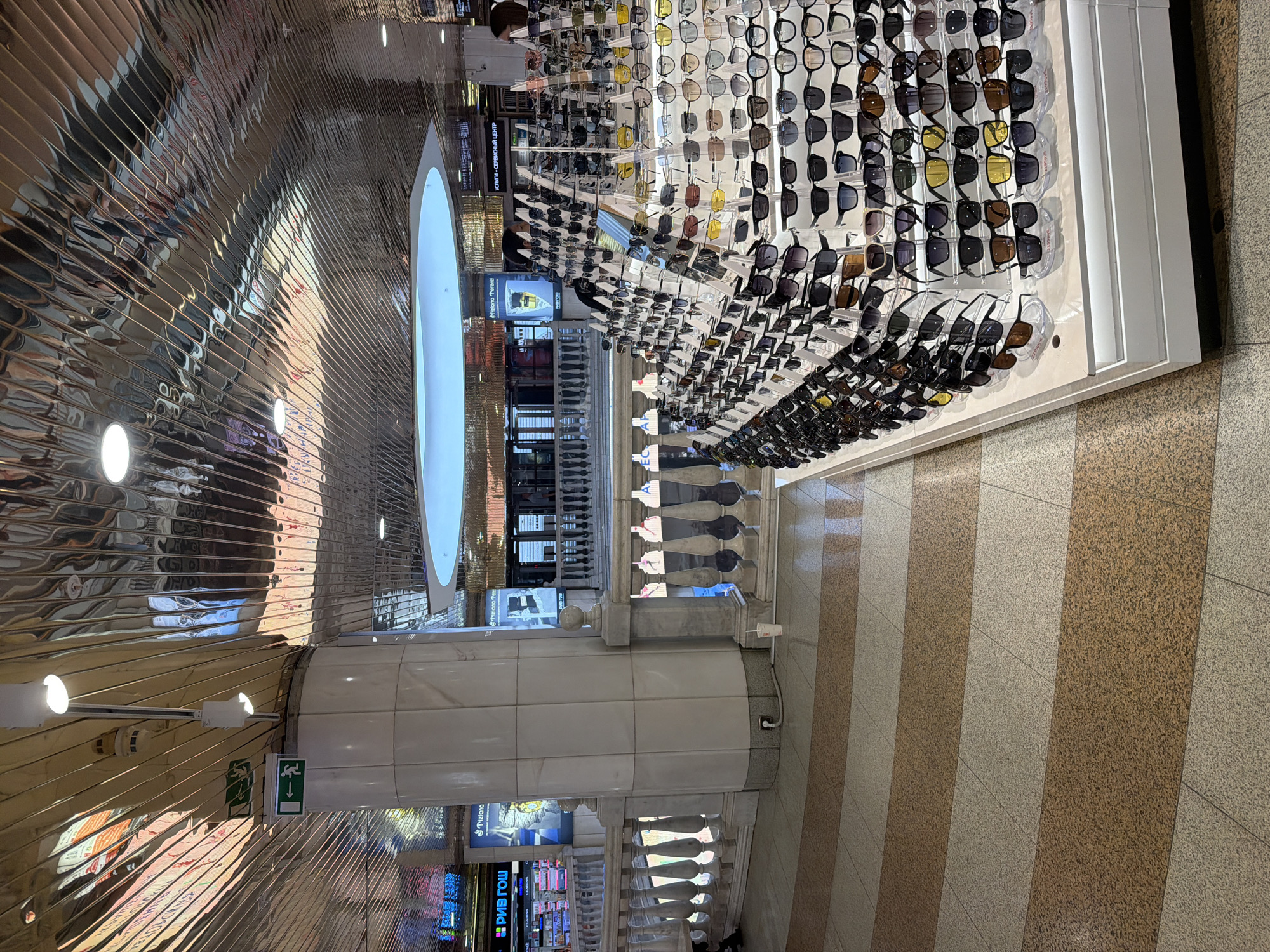 The image shows the interior of the TSUM department store in Moscow, Russia. The perspective is looking upward and slightly to the side at a multi-story retail space. Display shelves showcasing a wide variety of sunglasses line the upper level of the store. Below this display, mannequin torsos stand behind a decorative railing. Large pillars with intricate architectural details and display screens support the structure. The floor has a striped pattern of light and dark tiles. A reflective, textured ceiling adds to the visual complexity of the space. A large circular light fixture illuminates the area. Signage in Russian is visible on the lower levels.
