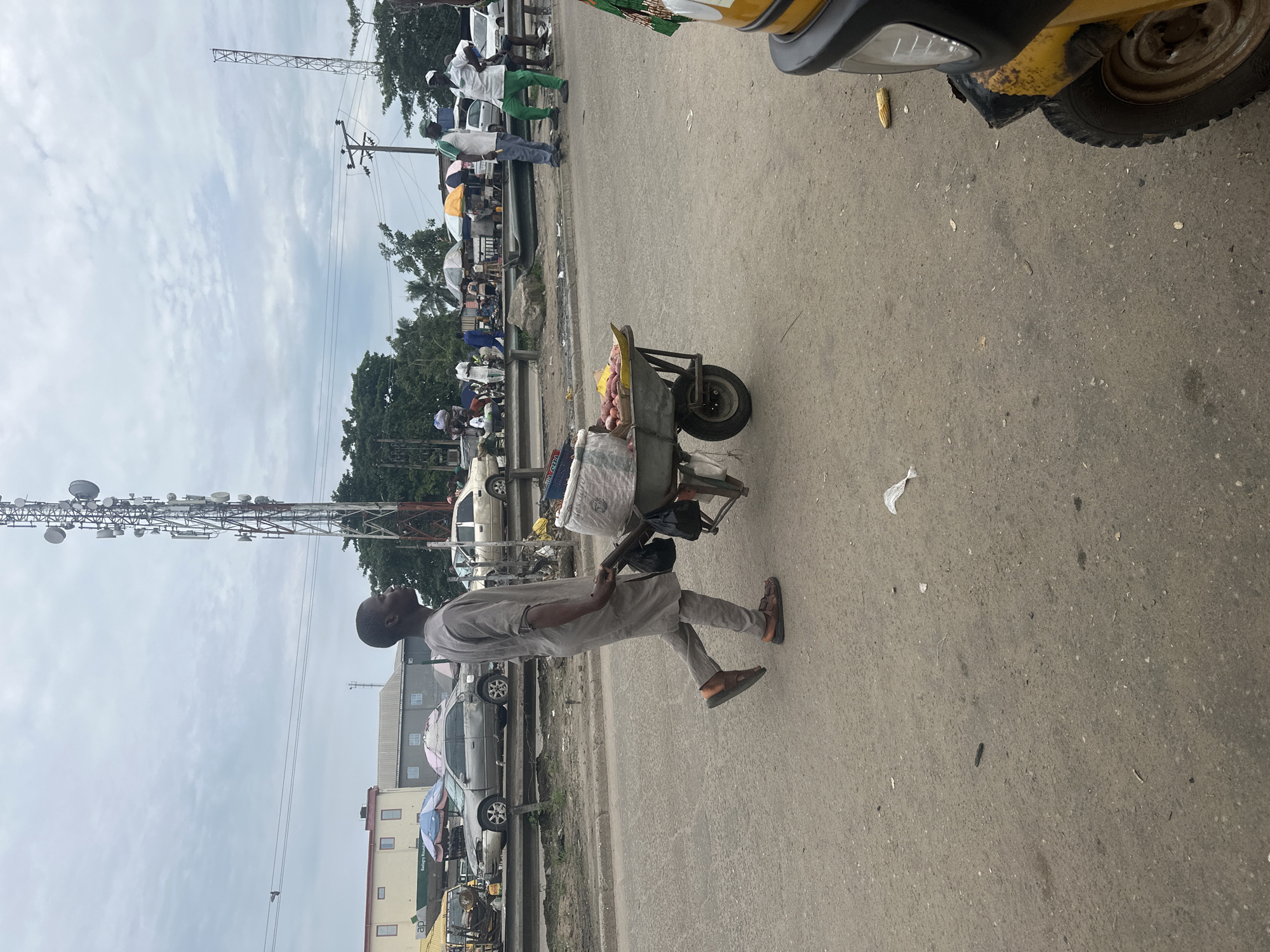 The image shows a man pushing a wheelbarrow loaded with goods, including what appear to be onions and bags of produce. He is wearing a light-colored, traditional outfit and sandals. The scene is set on an asphalt road, likely in a marketplace or a busy street in Badagry, Nigeria. In the background, there are various vehicles, including cars and a yellow motorized tricycle. Further in the distance, a large telecommunications tower is visible, along with trees and buildings. The sky is overcast with scattered clouds, suggesting it might be daytime with possibly impending rain. The overall atmosphere appears to be that of a bustling commercial area, with people engaged in daily activities. There are no discernible emotions clearly visible on the man's face, but the act of pushing a loaded wheelbarrow suggests he is working. There is some visible text on a green sign in the background, but it is too blurry to read.
