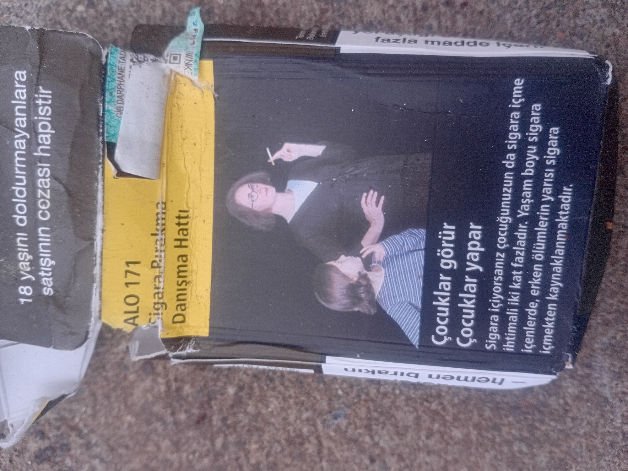 The image shows a close-up of a crumpled cigarette pack with a health warning. The cigarette pack is predominantly black with a yellow band at the top. The yellow band displays text in Turkish, including "ALO 171" and "Sigara Bırakma Danışma Hattı" which translates to "Quit Smoking Helpline." Below the yellow band, a photo depicts a woman smoking a cigarette and a child. The text on the pack also includes a health warning in Turkish that reads "Çocuklar görür, çocuklar yapar. Sigara içiyorsanız çocuğunuzun da sigara içme ihtimali iki kat fazladır. Yaşam boyu sigara içenlerde, erken ölümlerin yarısı sigara içmekten kaynaklanmaktadır." This translates to "Children see, children do. If you smoke, your child is twice as likely to smoke. Half of the early deaths in lifelong smokers are caused by smoking." A vertical text on the right side of the pack reads "fazla madde ig..." which is part of a legal notice. The cigarette pack appears to be lying on a rough, grey surface, possibly pavement or concrete, which might be located in Onikişubat, Turkey, given the context of Turkish text. The pack is damaged, with torn edges and creases, suggesting it has been handled or discarded.