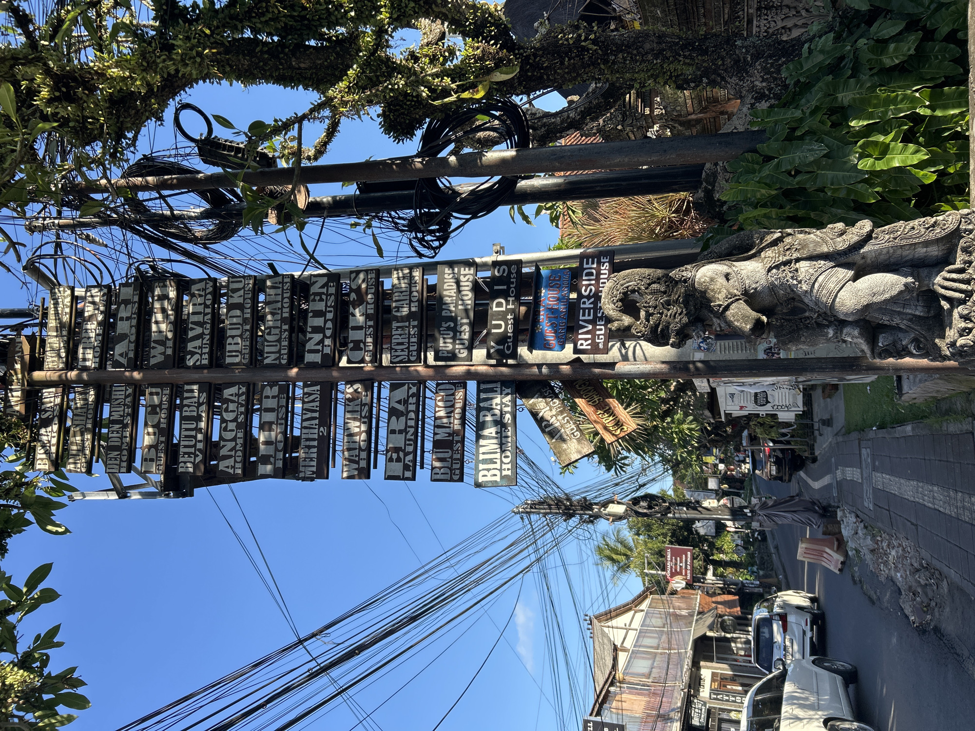 This is a photograph taken in the Ubud District of Indonesia. The image depicts a street scene filled with numerous signs hanging from a utility pole. The signs advertise various accommodations like guest houses, villas, and homestays, along with services such as laundry. Some prominent names visible on the signs include "Savira Bungalow," "Ubud Ku Guest House," "Betutu Bali Villas," "Inten House," "Secret Garden," "Riverside Guest House," and "Blu Mango Guest House." The street appears to be lined with buildings and trees, suggesting a bustling urban or tourist area. Overhead, a tangle of electrical wires crisscrosses the sky, typical of many Southeast Asian streets. A carved stone sculpture, possibly of a mythical creature or deity, is visible on the right side of the frame, adding a cultural element to the scene. The bright sunlight and clear blue sky indicate it is daytime, likely mid-morning or afternoon. The overall impression is of a vibrant and well-traveled area, bustling with commerce and accommodation options.