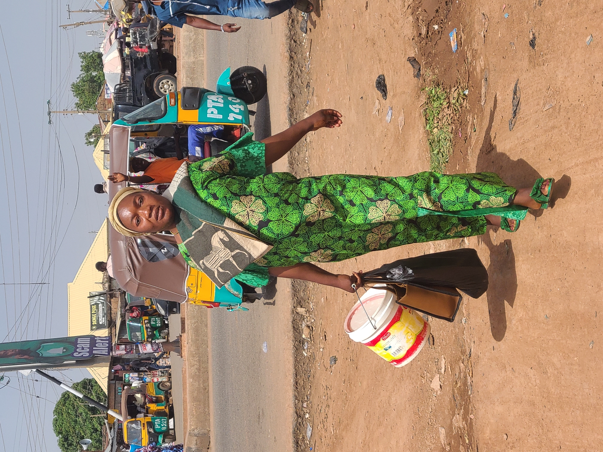 A woman stands on a dirt and asphalt surface near a street with several three-wheeled vehicles in the background. She wears a green patterned dress and a head covering, holding a bucket in one hand and a dark-colored bag in the other. The environment appears to be a busy street scene, potentially in a commercial area with visible signs and parked vehicles. The location is Jos, Nigeria, but is not explicitly shown but implicitly indicated by the style of transport and dress of the woman.
