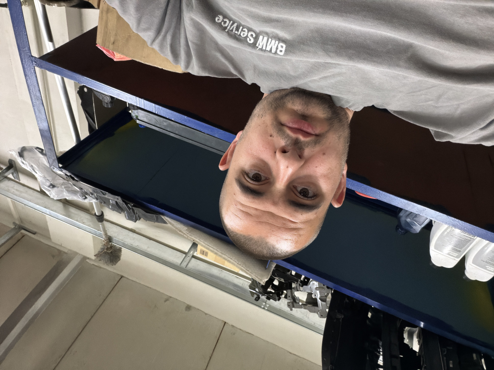 The image shows a man in a grey t-shirt with "BMW Service" printed on it, taking a selfie. He has a shaved head and is looking directly at the camera. The background appears to be a workshop or storage area, with shelves containing various items, including bottles, tools, and metal parts. The shelves are painted blue and red. Above the shelves, the ceiling has exposed metal beams and panels. A duster with a long handle is leaning against the top shelf. The overall lighting suggests an indoor environment. The man's facial features indicate he is of adult age. The specific location in Burgas, Bulgaria is not discernible from the image itself.