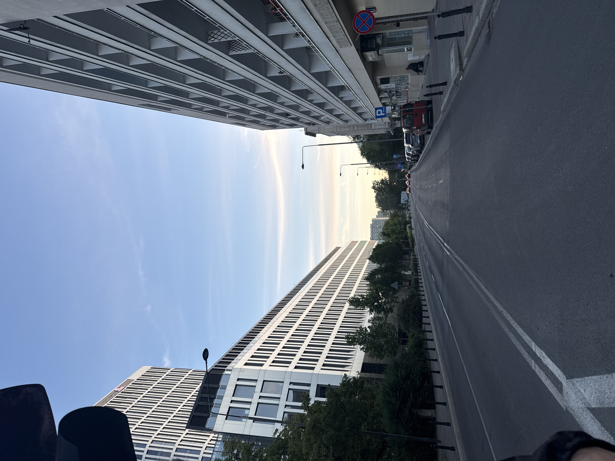 The image depicts a street scene in an urban environment. On the left side of the street, a modern, multi-story building with a white facade and numerous dark-framed windows dominates the view. Trees line the sidewalk in front of this building. On the right side, another building with a lighter colored facade features a repetitive pattern of balconies or recessed windows. The street itself is asphalt with white lane markings. Several cars are parked along the right side of the street. A few streetlights are visible. In the distance, more buildings and cars can be seen. The sky is clear with some wispy clouds and is transitioning towards evening light. A traffic signal is partially visible in the top left corner of the image. A person is seated on a ledge in front of the building on the right.