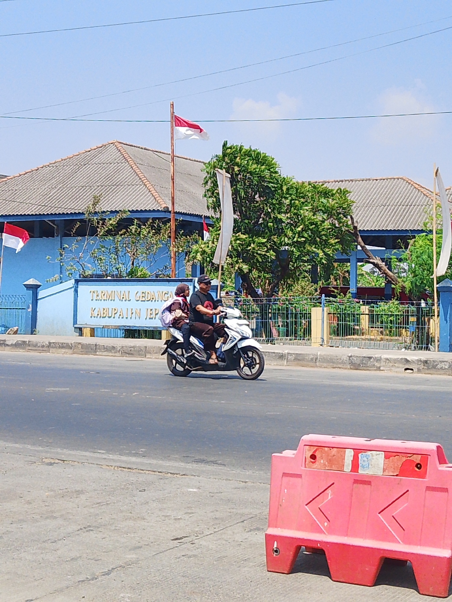 A medium shot shows a sunny outdoor scene with a blue building labeled "TERMINAL GEDANGAN KABUPATEN JEP" in white letters. Indonesian flags, red and white, are visible flying from poles in front of the building and to the left. A white motorcycle carries two people, a man in a black t-shirt and sunglasses, and a younger person in a brown jacket and hijab, as they ride on a paved road. A red traffic barrier is positioned in the foreground, on the right side of the image. Trees and a fence are visible behind the motorcycle, near the building. The sky is clear blue with some thin power lines running across it.