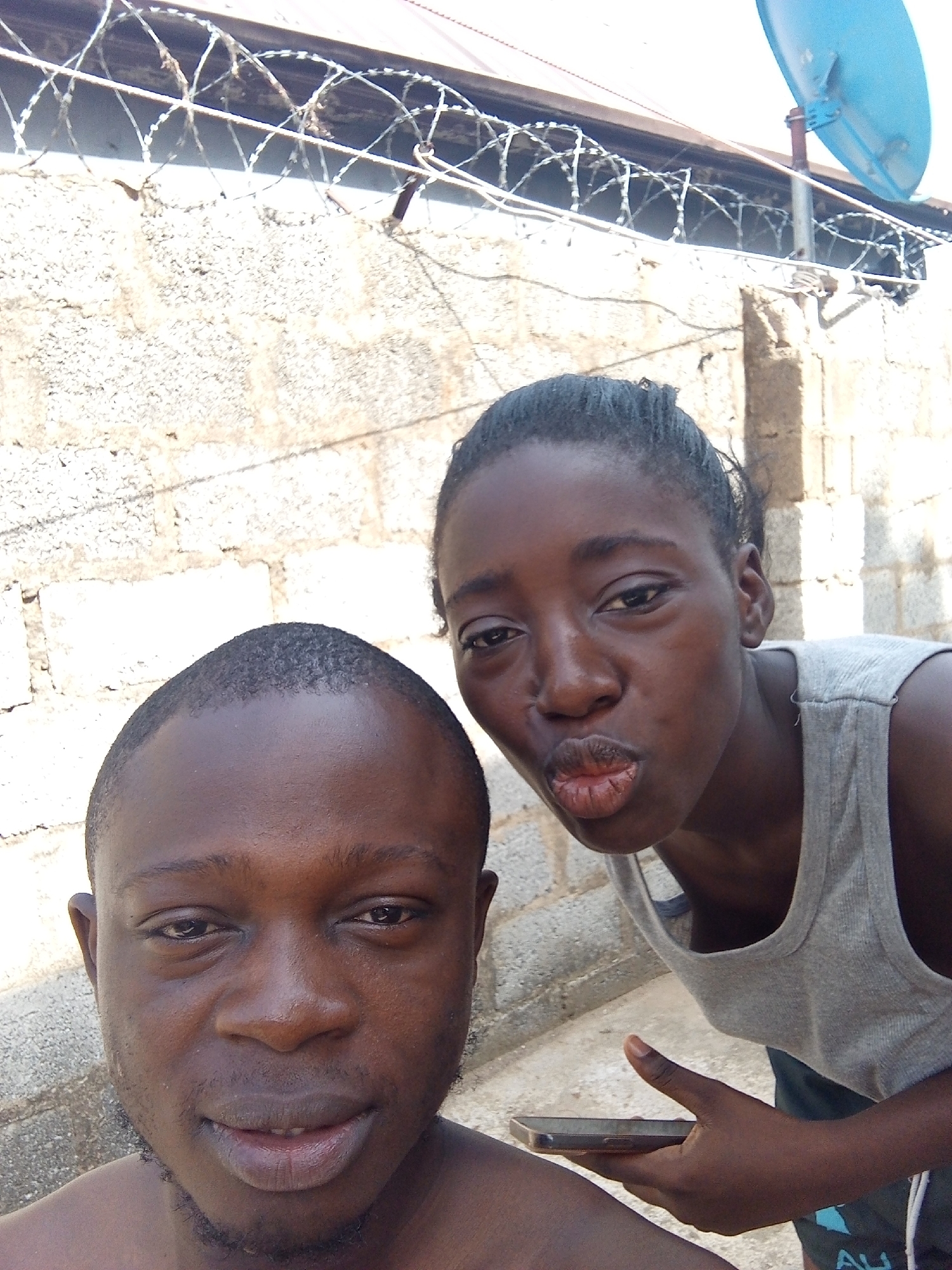 Two people, a male and a female, are in the foreground of the image. The male is shirtless and positioned closer to the camera. The female is wearing a gray tank top and is making a kissing face. She is holding a mobile phone. The background includes a concrete block wall topped with barbed wire, and a satellite dish is visible above the wall. The image appears to be taken outdoors during daylight. There is no direct evidence of location. There is no definitive information to place the location in Abuja, Nigeria.