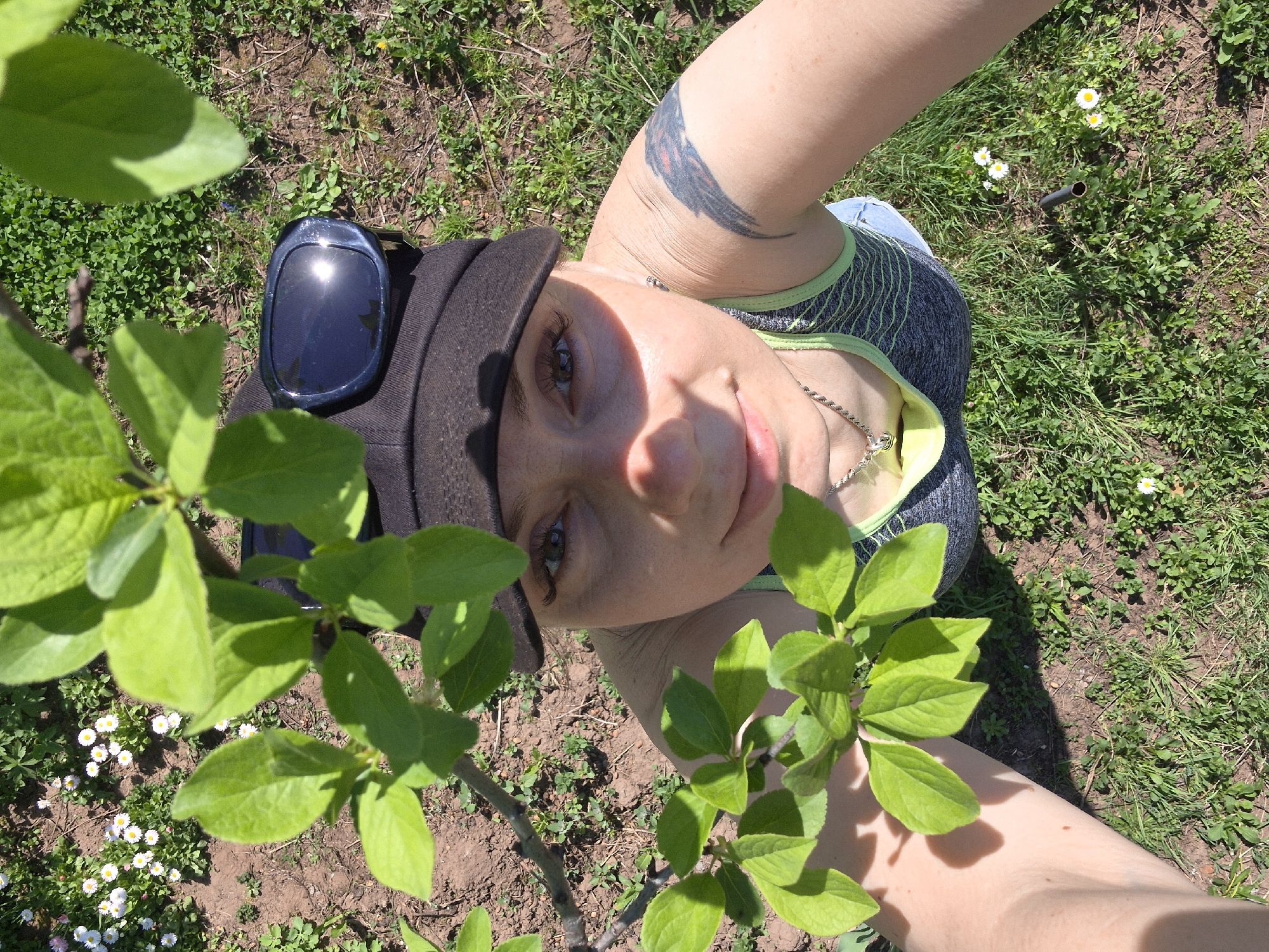 A woman wearing a black cap with sunglasses on top is lying on the ground in an area with grass, small white flowers, and bare dirt patches. She is wearing a gray and green top and a silver chain necklace. A tree branch with green leaves is positioned between the viewer and the woman's face. Her right arm, which has a tattoo, is raised. The scene appears to be outside in daylight. The specific location is described as the city of Chaikovsky, Russia.