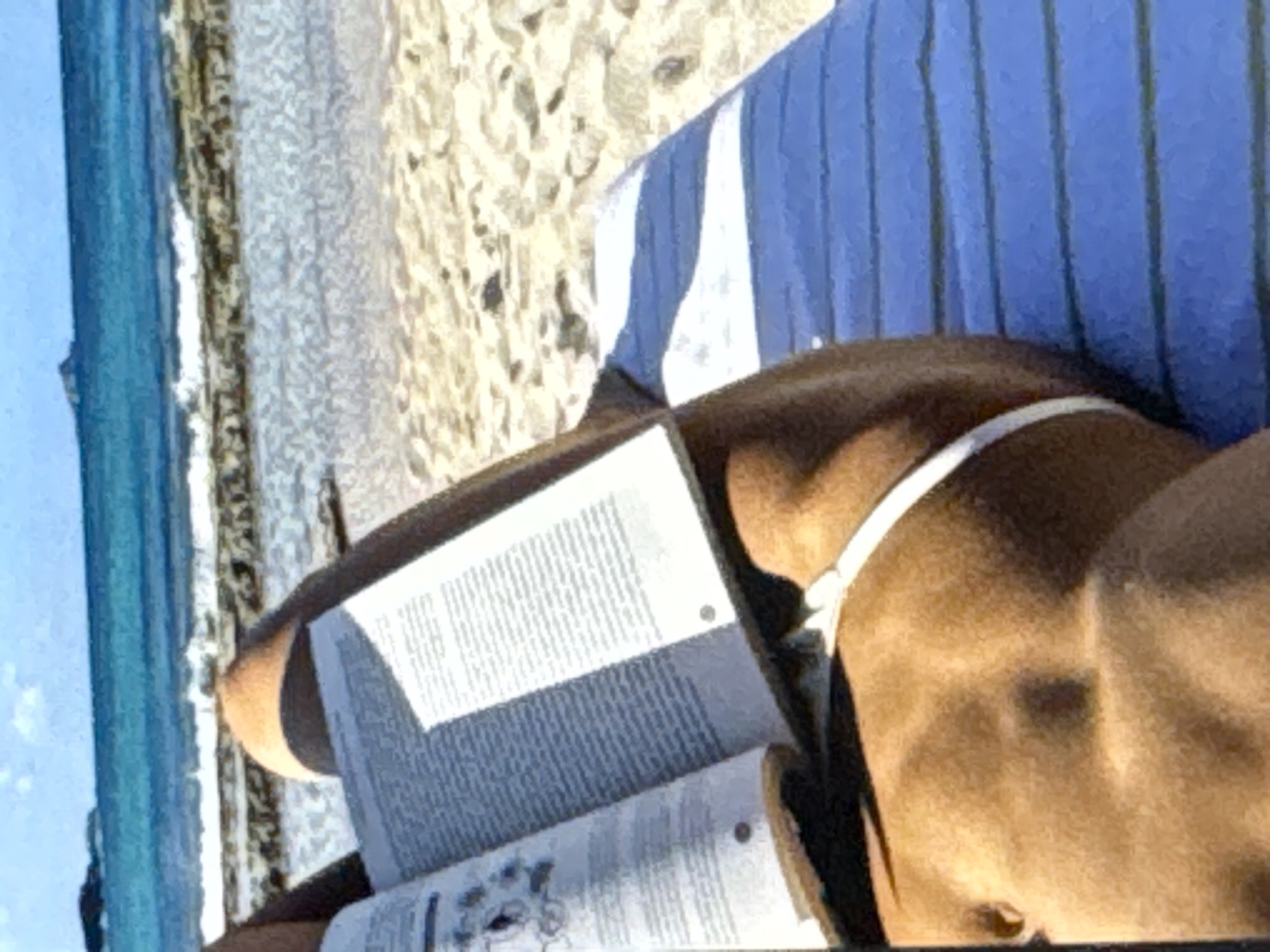 This is a close-up, vertical, eye-level shot of a person lying on a beach. The person is wearing a blue and white striped shirt and a white bikini bottom. They are holding an open book, with text visible on the pages. The book is casting a shadow on the person's chest and the sand. The background shows a sandy beach with the turquoise water of the ocean on the left side of the frame. The sky is a bright, clear blue. The image appears to have been taken on a sunny day. The scene suggests a relaxed atmosphere, with the person enjoying reading on the beach.