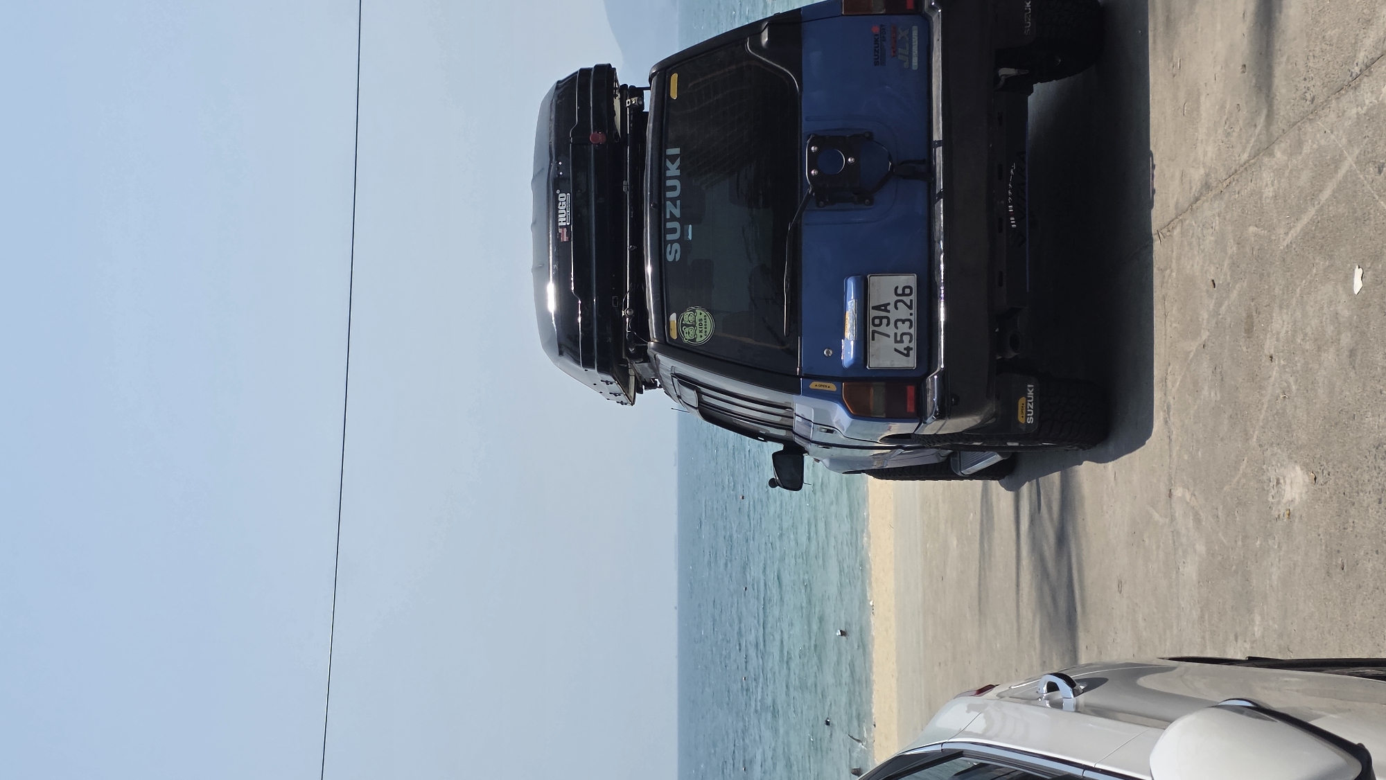 The image captures a bright, sunny day near the beach in Nha Trang, Vietnam. A light blue Suzuki SUV is parked on a concrete surface. The SUV has a black roof cargo carrier. The license plate number is "79A 453.26." The SUV is parked perpendicular to the beach, which is visible in the background with the ocean stretching into the distance. A partial view of a white car is visible in the foreground. Overhead power lines are visible.