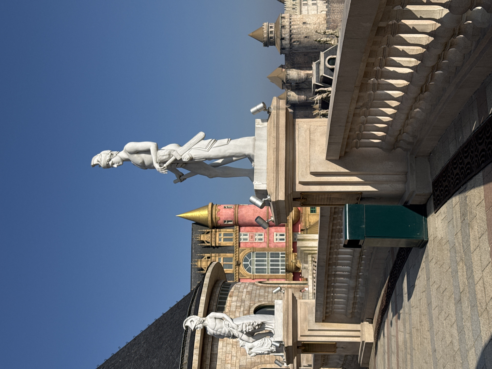 The image captures an outdoor architectural scene at Sun World Ba Na Hills, Đà Nẵng, Vietnam, under a clear, cloudless blue sky.
In the immediate foreground, a patterned stone-tiled walkway is visible, featuring a dark green, rectangular trash receptacle positioned to the right. A light-colored stone balustrade with multiple balusters extends horizontally across the mid-ground, casting distinct shadows from the strong sunlight.
Two white marble statues of male figures in classical style stand on elevated pedestals behind the balustrade. The more prominent central statue depicts a muscular male figure wearing a helmet, holding a sword or similar object in his hands. To the left, a second, smaller statue portrays a male figure holding a severed head, positioned in front of a curved, light-colored building with a dark-tiled roof. Each statue's pedestal is equipped with multiple silver-colored spotlights.
In the mid-ground, a vibrant pink multi-story building with ornate windows, dark roofs, and a prominent golden-domed turret is visible. A small, indistinct human figure is partially visible on a balcony of this pink building. Further in the background, to the right, a sprawling stone fortress or castle complex with multiple circular towers, battlements, and various smaller statues on its rooftops extends towards the horizon.