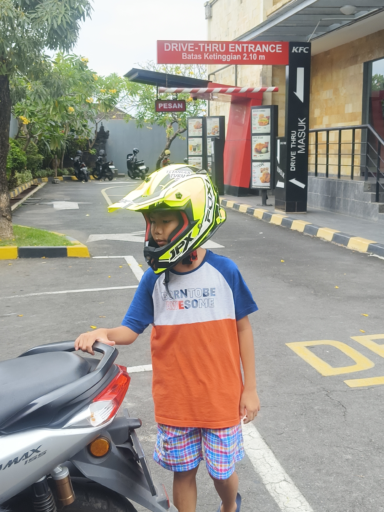A young boy wearing a yellow and black motocross helmet and a t-shirt with the text "BORN TO BE AWESOME" stands next to a silver scooter at a KFC drive-thru in Denpasar, Indonesia. The drive-thru entrance sign reads "DRIVE-THRU ENTRANCE" and "Batas Ketinggian 2.10 m". A menu board displays food items. Several motorcycles are parked nearby, and lush green trees with yellow flowers are visible. The ground is asphalt with yellow painted lines.