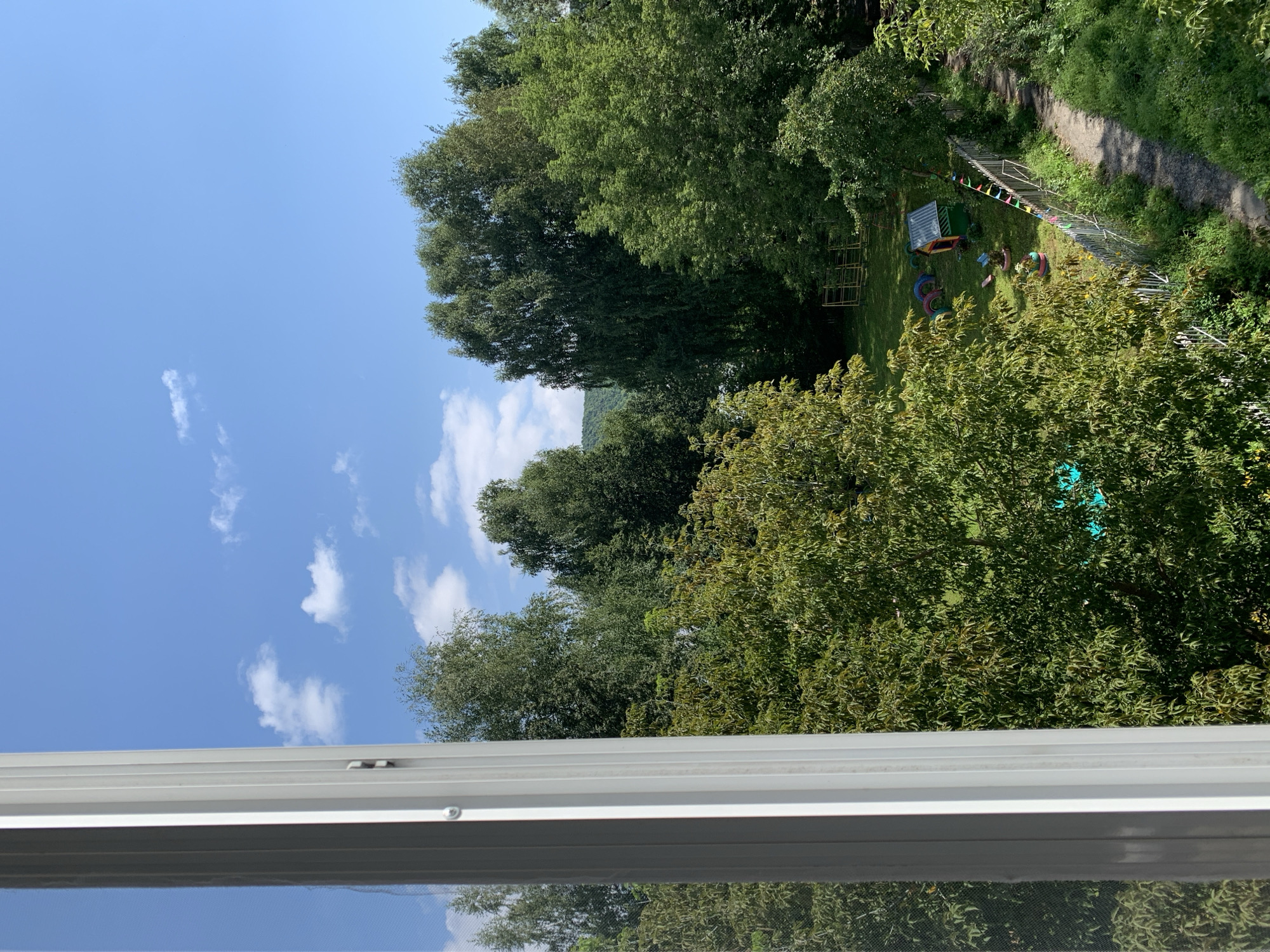 The image shows a view from a window of a yard with trees, a playground, and a path. The sky is blue with scattered white clouds. In the foreground, a white window frame is visible on the left. In the yard, there are several trees of varying heights, a small wooden playhouse with a green roof, a set of swings, and some colorful tire sculptures. A gravel path runs along the right side of the yard, bordered by a metal fence decorated with colorful flags. In the background, there are more trees and a distant hill. The overall impression is one of a sunny day in a residential area. This scene is located in the city of Asha, Russia.