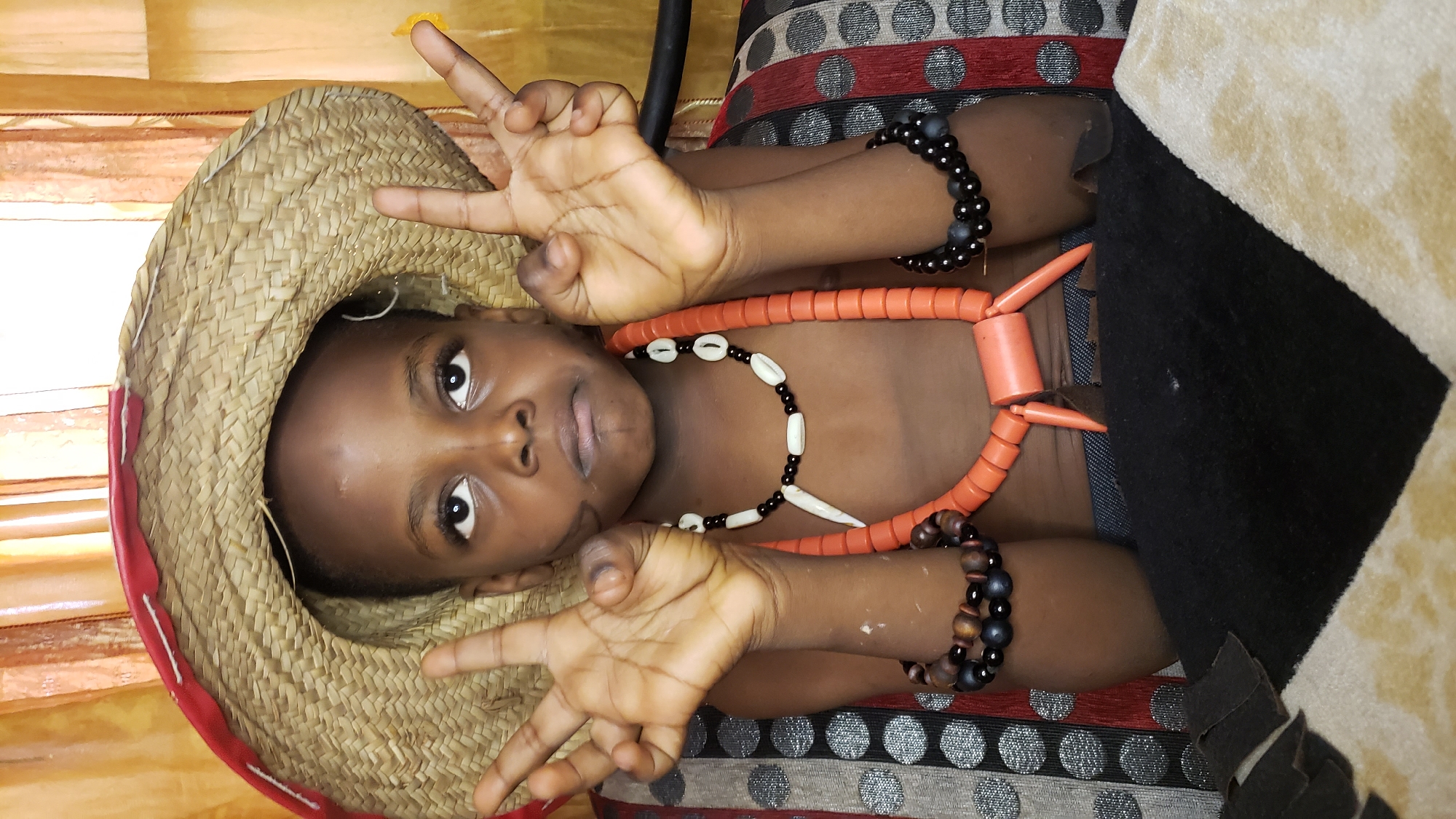A young Black child, wearing a straw hat with a red brim, faces the camera with a slightly pouty expression. The child's arms are raised, and both hands are held up, displaying the peace sign with their index and middle fingers extended. The child is adorned with several necklaces, including a prominent orange beaded necklace with pointed accents and a smaller black and white beaded necklace. Bracelets are visible on both wrists: a dark beaded one on the left and a multi-colored wooden and beaded one on the right. The child is seated, with a patterned couch visible behind them and a dark fabric in the foreground. The background consists of sheer, orange-toned curtains. The scene appears to be indoors, likely within a residential setting in Sabon Tasha, Nigeria.