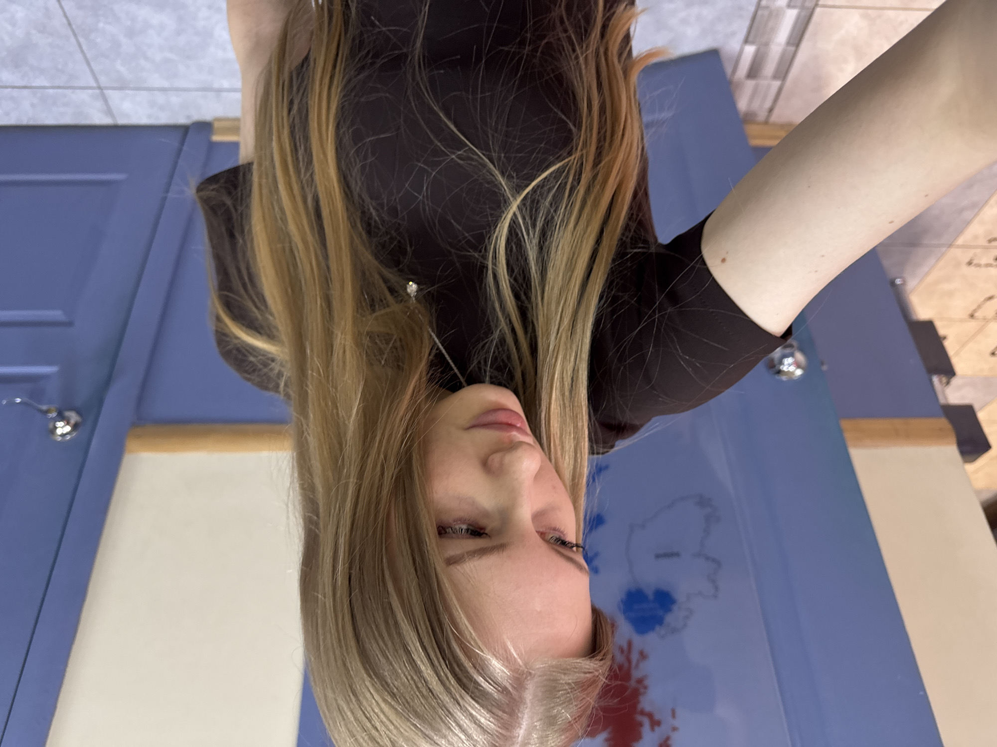 The image is a selfie taken by a young woman with long, blonde hair wearing a dark brown top. Her face is slightly angled towards the camera. In the background, there is a blue surface with a map of Russia, featuring some red and blue areas. To the left of the map is a blue cabinet with a chrome handle. Beyond the map and cabinet, there is a glimpse of a white wall with a tile pattern. It is not possible to confirm the location in the city of Perm, Russia without additional information.