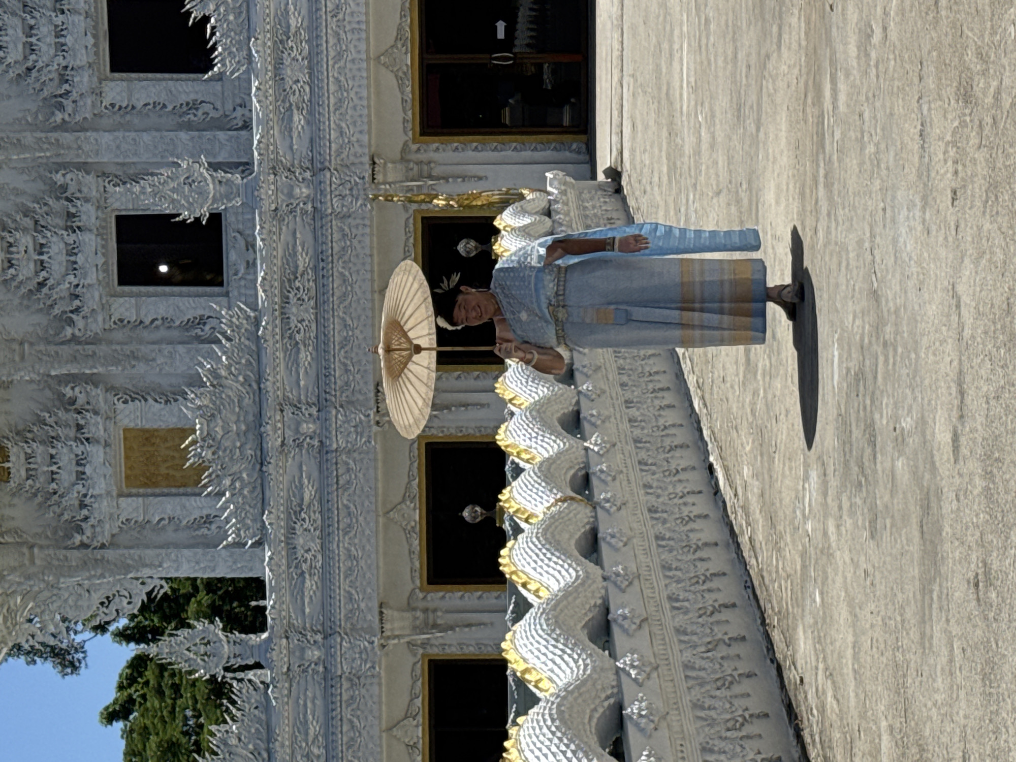 A full shot shows a woman standing in front of a white temple with ornate decorations and dark windows. She is wearing a traditional light blue and yellow outfit, a floral headpiece, and holding a light brown umbrella. She has her right hand on the umbrella and is smiling. The temple has a low wall with a decorative dragon-like pattern in white and gold. The ground is a light grey concrete walkway with a shadow of the woman cast on it. The sky is clear and blue.