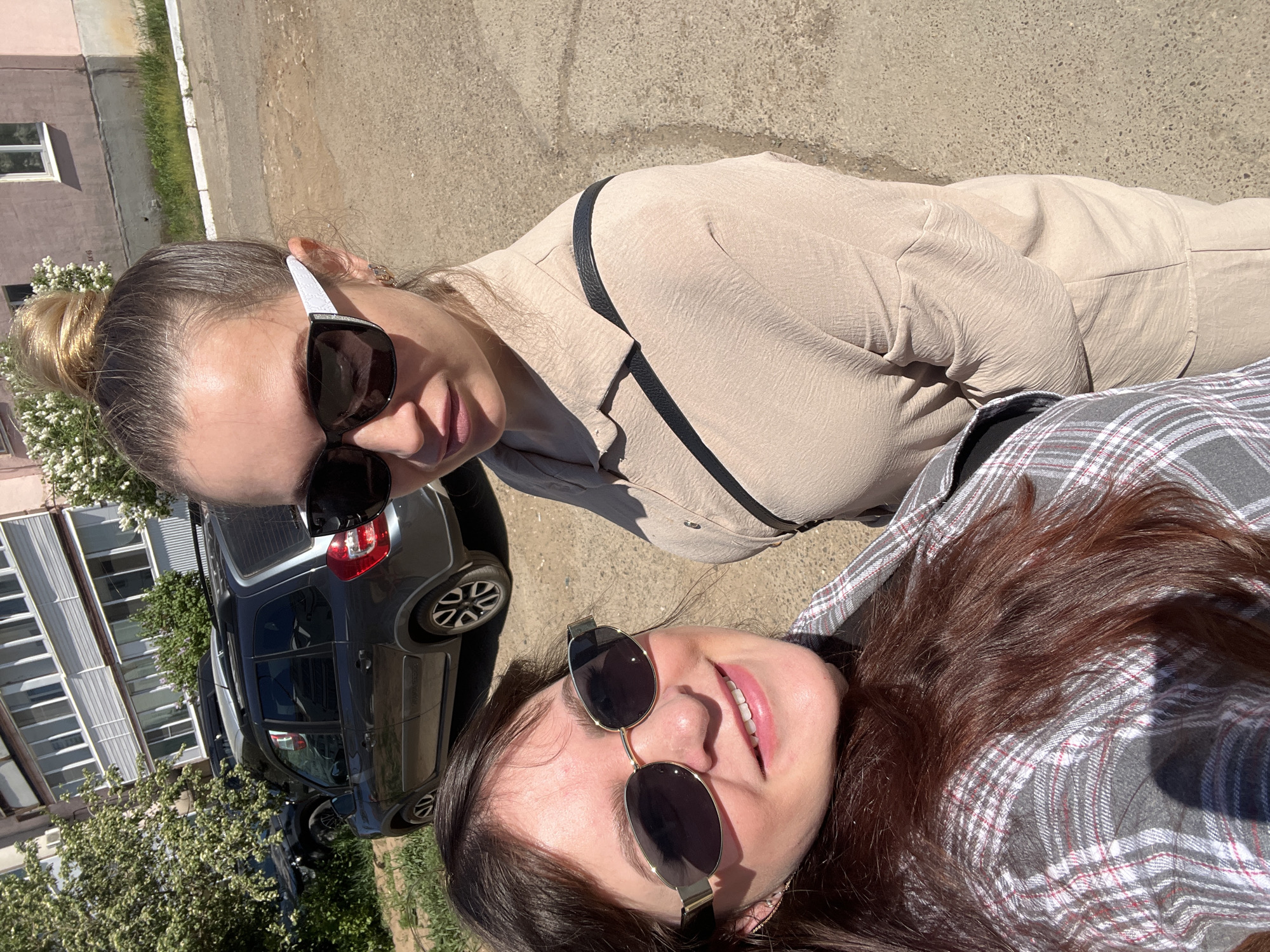 Two women are pictured in an outdoor selfie, possibly taken on a street. The woman on the top, closer to the camera, has blonde hair pulled back in a bun and is wearing sunglasses and a light beige, button-down shirt or jacket. The woman underneath her has dark brown hair and is wearing sunglasses and a gray and white plaid shirt. Behind them, a gray hatchback car is parked. In the background is a multi-story apartment building with white windows and a small tree with white flowers. The location appears to be a paved area. The city mentioned is Noviy, Russia.