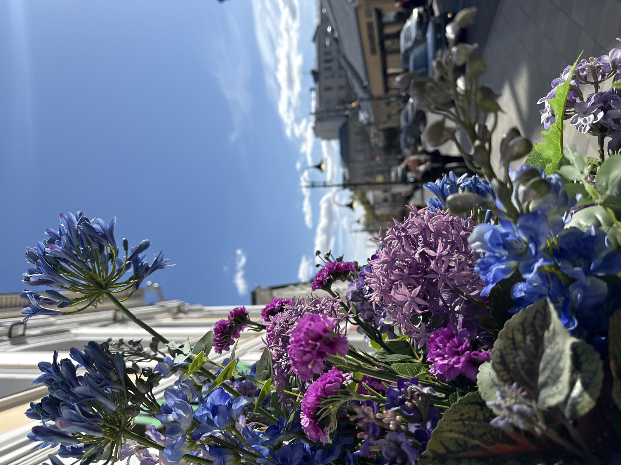 The image shows a bouquet of artificial blue and purple flowers in the foreground. The background features a street scene with buildings and cars, partially obscured by the flowers. The sky is bright blue with some clouds. The location is Moscow, Russia, identifiable from the architectural style and streetscape. People are visible in the background, walking along the street.