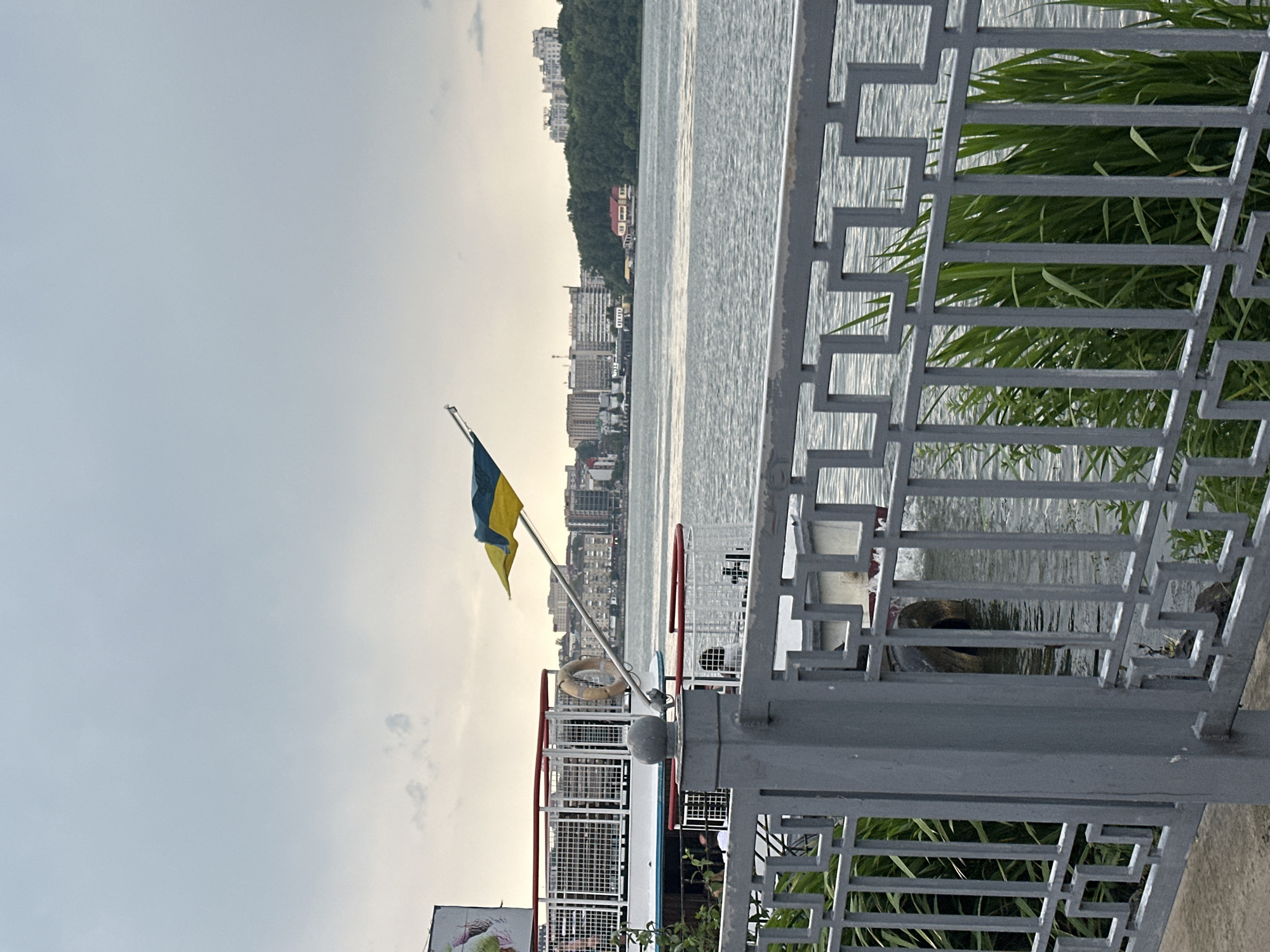 The image shows a view of Ternopil Lake in Ternopil, Ukraine, on an overcast day. In the foreground, a gray metal fence with a geometric pattern runs horizontally across the frame. Behind the fence, lush green reeds and grass grow along the water's edge.
The middle ground features a white boat with red trim docked at a pier. A Ukrainian flag, a bicolor of blue and yellow, is prominently displayed on a flagpole attached to the boat. A life preserver ring is visible hanging on the side of the boat. Several people appear to be on the boat, though their details are not clear.
In the background, across the lake, a cityscape unfolds with numerous multi-story buildings. Beyond the buildings, a densely forested hill rises, adding a natural element to the urban landscape. The lake's surface is rippled, suggesting a slight breeze. The sky is a muted gray, typical of cloudy weather.