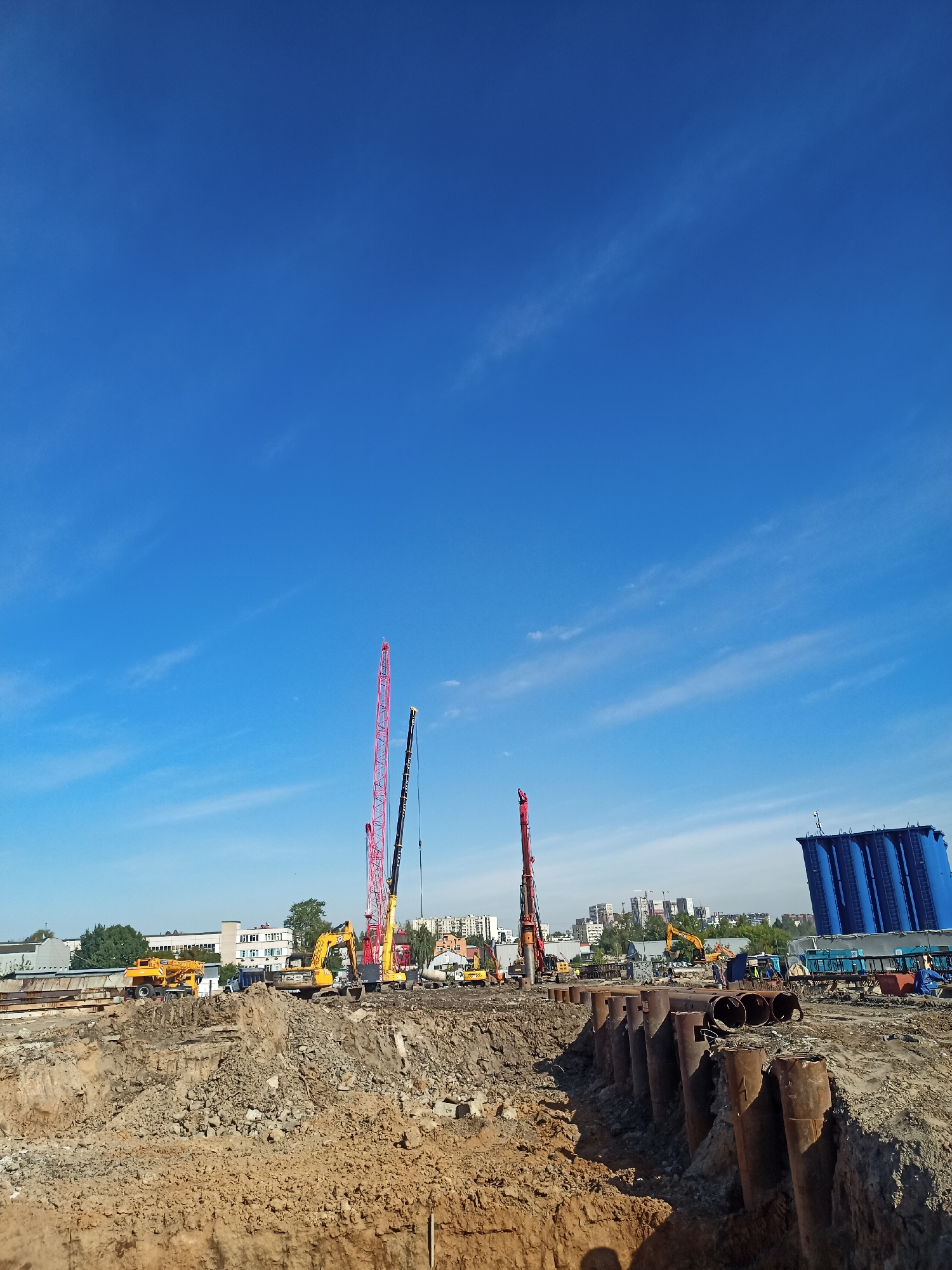 The image depicts an active construction site under a clear blue sky with wispy clouds. The foreground is dominated by a deep excavation filled with loose soil and rocks, with large, rusty metal pipes lining one side of the trench. In the mid-ground, a variety of heavy machinery is at work, including yellow excavators and cranes, one of which is a tall red lattice boom crane. In the background, buildings of various heights, including residential apartment blocks and industrial structures, form a city skyline. A large blue structure, possibly a storage tank or industrial equipment, stands prominently on the right side of the image. Several construction workers, dressed in safety vests, are visible near the machinery. The scene suggests ongoing infrastructure development or building construction within an urban environment.