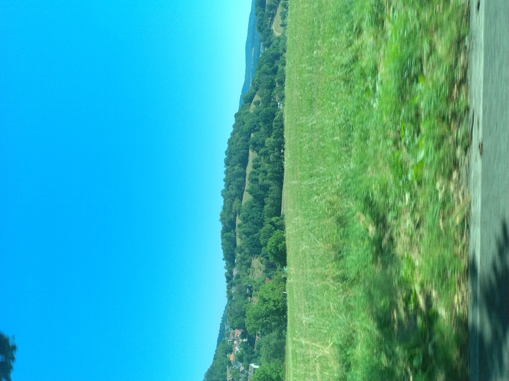 The image depicts a bright, clear blue sky above a lush, green hilly landscape. In the mid-ground, a village with red-roofed houses is nestled among trees on a slope. The foreground is dominated by a blurred field of green grass, suggesting motion or a shallow depth of field. The overall scene is one of a rural environment under a sunny sky. The location is identified as Brloh, Czechia.
