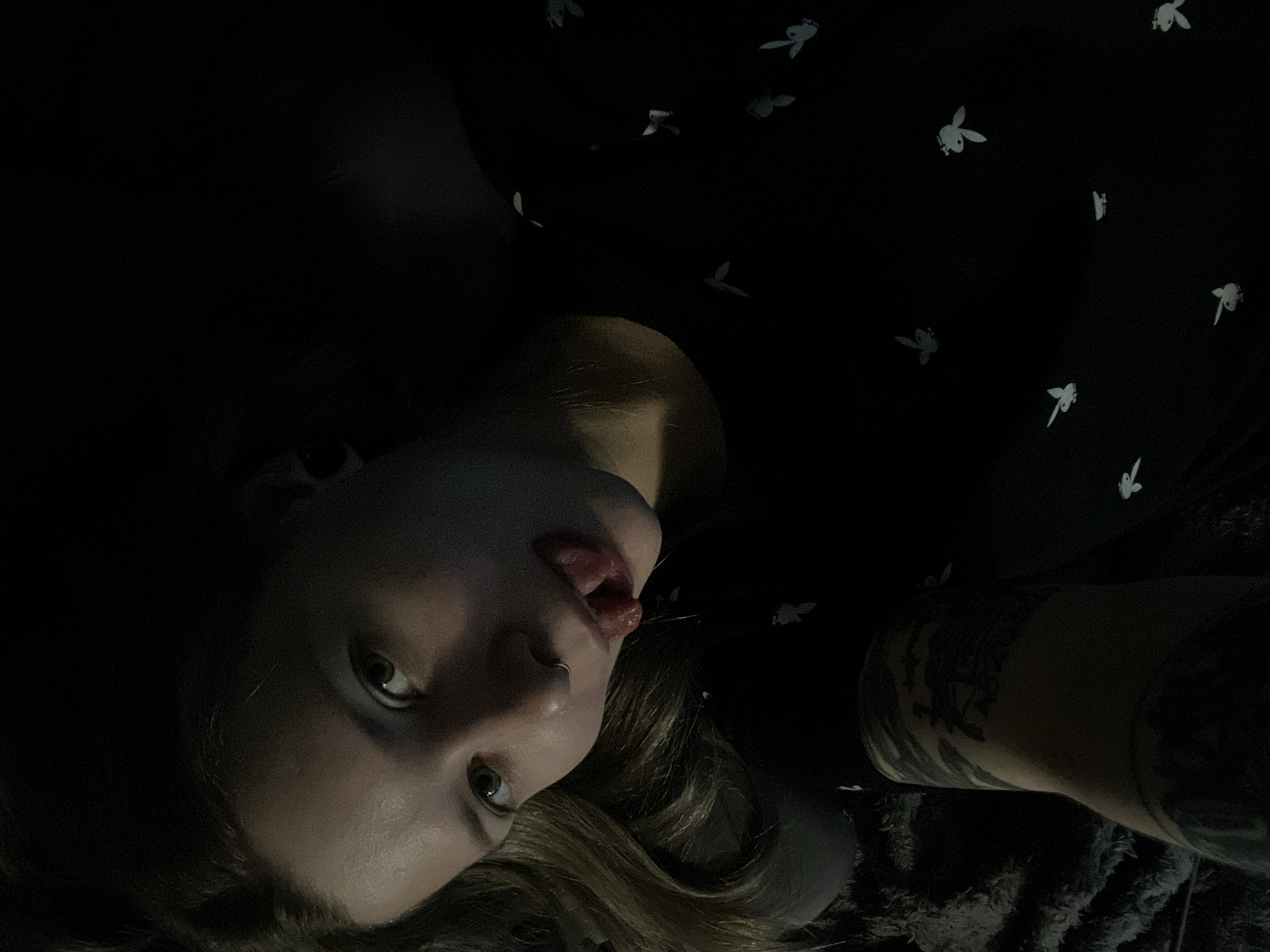 A woman is photographed in a low-lit environment. She is lying down and facing the camera. Her tongue is sticking out. She has a nose ring and visible tattoos on her arm. She is wearing a black shirt with a repeating Playboy bunny pattern. The background is dark. There is no evidence to suggest that the location is Khimki, Russia.