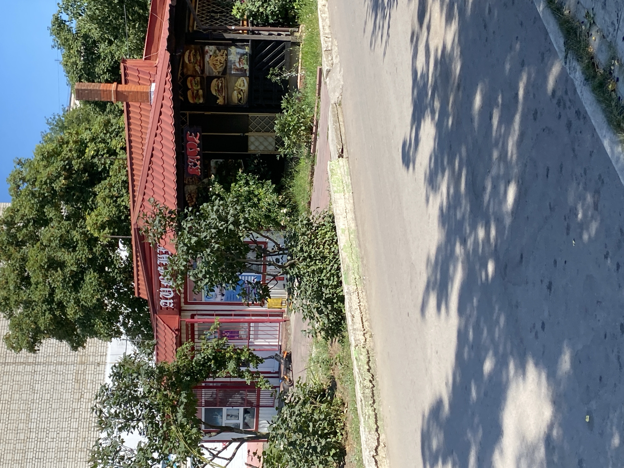 The image captures a street view in Chornomorsk, Ukraine, on a bright, sunny day. The foreground is dominated by an asphalt road with distinct shadows of trees cast across it. A concrete curb, partially painted green, lines the edge of the road. Behind the curb is a paved walkway with some grass and small bushes.
The main subject of the image is a small commercial building with a red corrugated metal roof. The building appears to be a cafe or food establishment, as indicated by images of burgers and fries displayed on its facade. The word "КАФЕ" is visible on a sign above the entrance. To the left of the cafe, there is another section of the building with a red-framed glass front, possibly a kiosk or shop. A yellow object, possibly a trash bin, is visible next to this section.
In front of the buildings, a few trees with green foliage provide shade. An electric scooter is parked on the paved area in front of the buildings. The background features a building constructed of light-colored brick and more lush green trees, with a clear blue sky above. A red brick chimney rises from the roof of the cafe. The overall scene depicts a typical urban commercial area on a clear day.