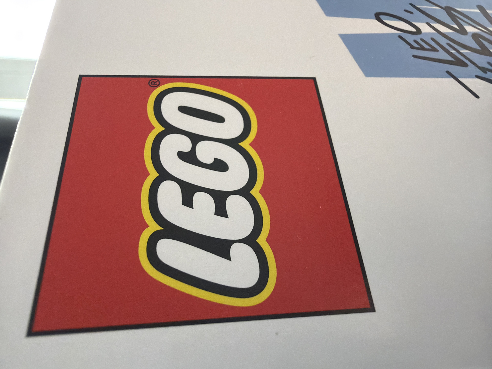 The image shows a close-up of a white box with the LEGO logo prominently displayed on its red surface. The LEGO logo is a red rectangle with the word "LEGO" in white, outlined in yellow and black. A small registered trademark symbol (®) is visible in the upper right corner of the red rectangle. The box is positioned at an angle, with part of a light-colored wall visible behind it. To the right of the LEGO logo, a portion of another design featuring blue and white stripes with stylized black lettering, potentially a name or word, is visible. The lighting suggests an indoor environment, possibly illuminated by natural light from a window. There are no people or specific city locations visible in the image. The image is a static shot of product packaging.