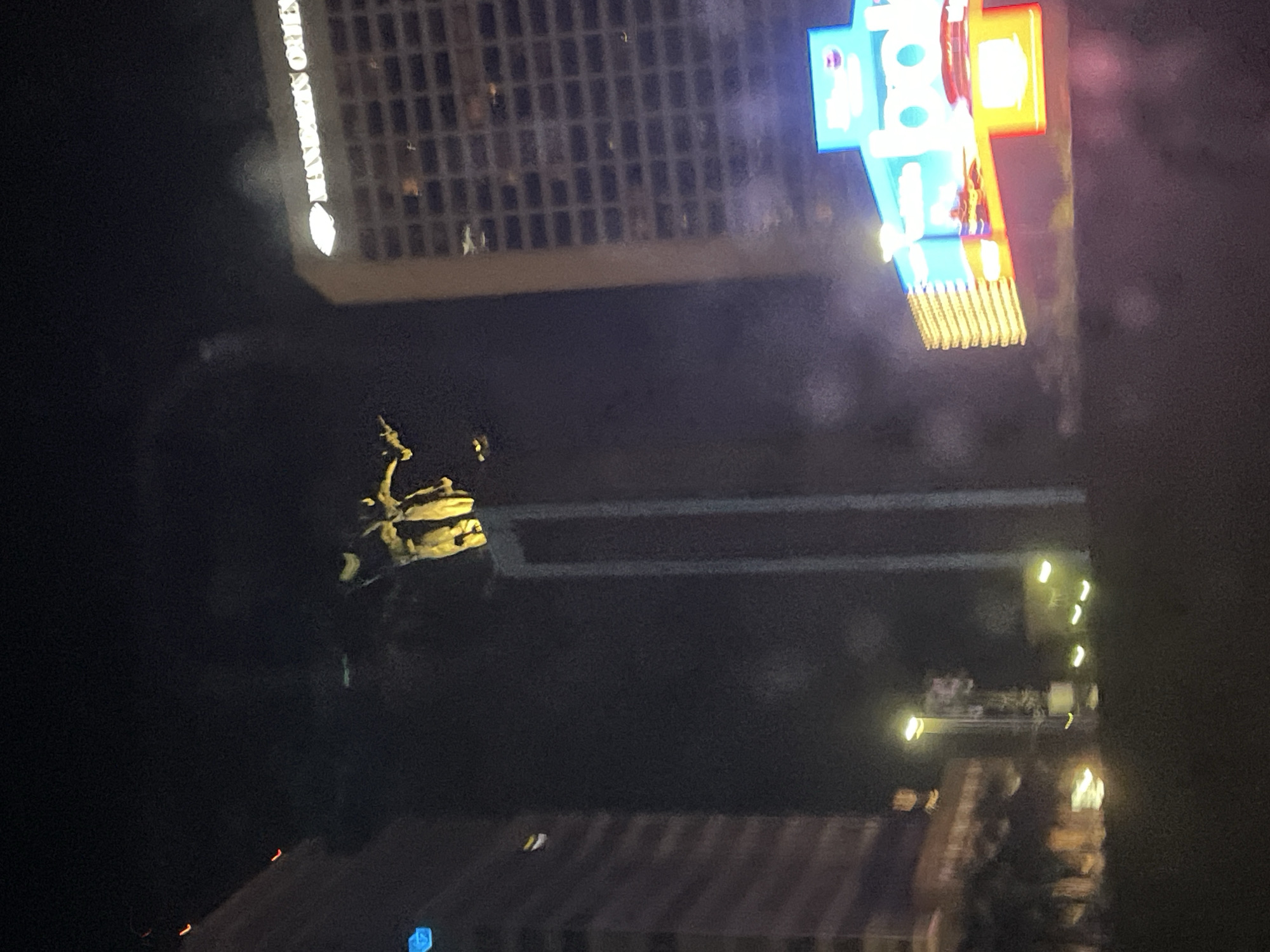 The image shows a night scene in Central Jakarta, Indonesia. A statue depicting a man on horseback is illuminated, with office buildings and a bright electronic sign in the background. Other city lights are visible in the distance.