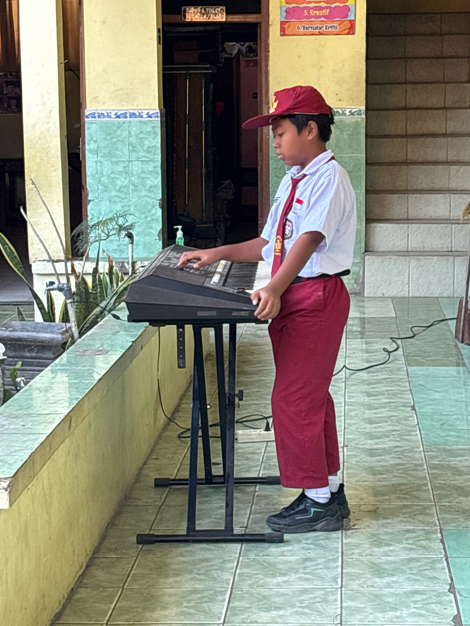 A young boy wearing a red and white school uniform, including a red cap, stands and plays a keyboard. The keyboard is black and rests on a black metal stand. The boy is positioned in profile, facing right, with his hands on the keys. He appears to be in a school or institutional setting, with tiled flooring and walls. Behind him, there are plants and a doorway. A sign above the doorway reads "DAPUR & TOILET" which translates to "Kitchen & Toilet." The overall scene suggests an outdoor or semi-outdoor area within a building complex in Tirtomoyo, Indonesia, likely a school.
