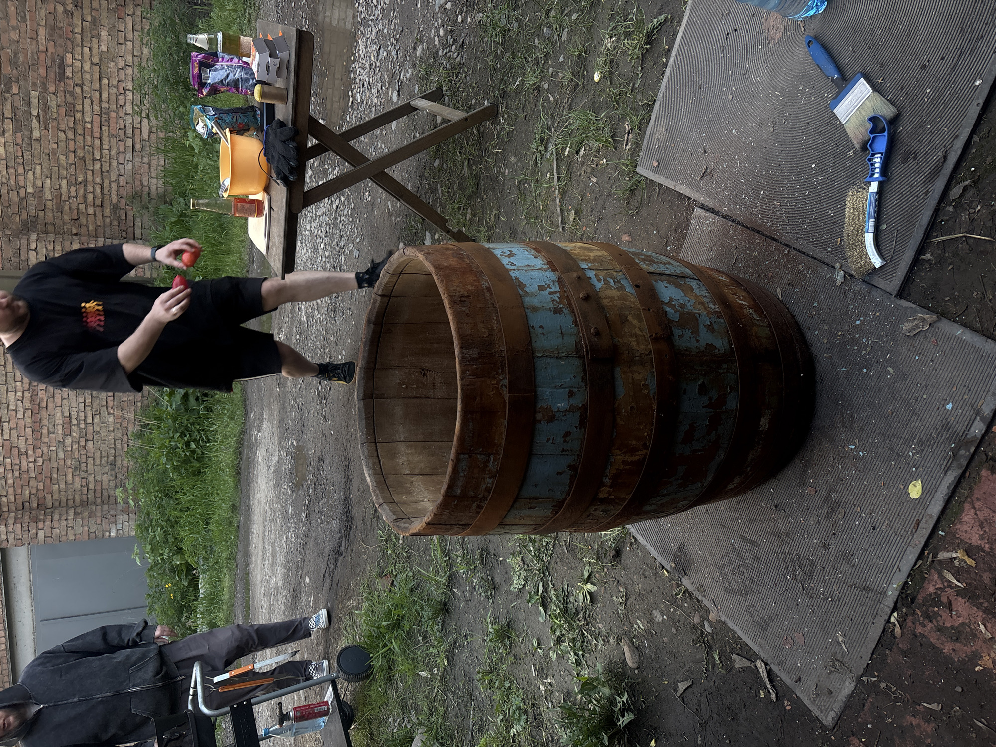 The image captures an outdoor scene in Krasnoyarsk, Russia. A weathered wooden barrel, banded with rusting metal, sits on the ground. A man in a black t-shirt and shorts stands next to the barrel, juggling two red objects. Adjacent to the barrel are gray rubber mats and a folding table laden with assorted items: a yellow bucket, food items in packaging, gloves, and bottles. The ground surrounding these objects is a mixture of dirt, gravel, and sparse green vegetation. In the background, there is a brick building wall, part of a grill and person. The overall lighting suggests an overcast day.
