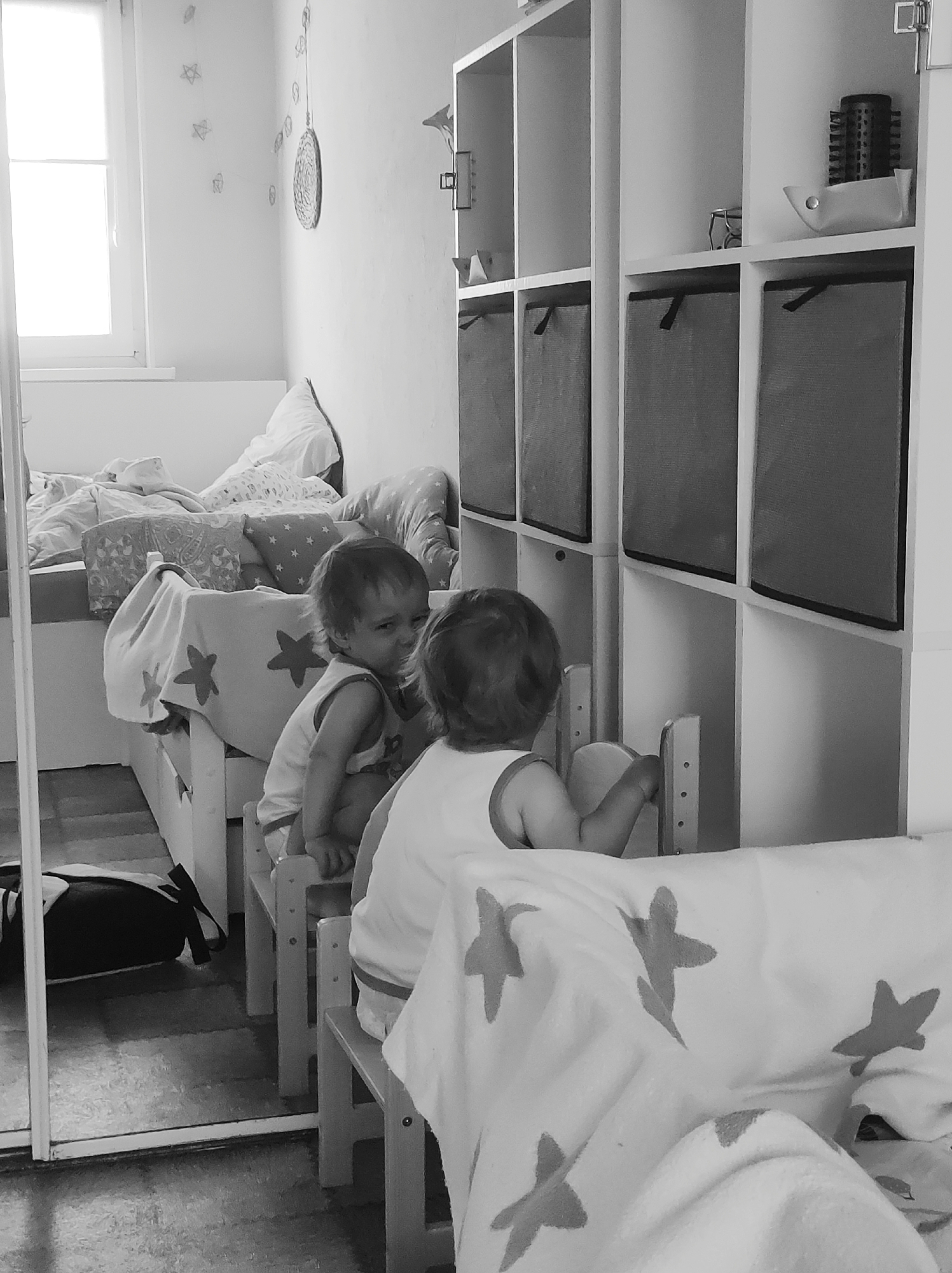 A black and white photograph captures two toddlers in what appears to be a child's bedroom or play area, potentially in Chaikovsky, Russia. One toddler is seated on a small wooden chair with their back mostly to the viewer, facing a white modular shelving unit filled with cubbies. The upper shelves hold a hairbrush and a small pouch, while the lower cubbies contain dark fabric storage bins.
The second toddler is visible in a floor-length mirror positioned on the left side of the room, also sitting on a small wooden chair. This child looks towards the first, exhibiting a focused or pensive expression. The mirror also reflects part of the floor and a dark backpack.
Natural light floods the room from a window on the far left, suggesting it is daytime. The walls are light-colored and decorated with a garland of stars and a circular hanging ornament. Behind the mirrored reflection of the child, a bed is visible with bedding patterned with stars and several pillows. The scene conveys a quiet moment of interaction between the two children. No text is visible in the image.