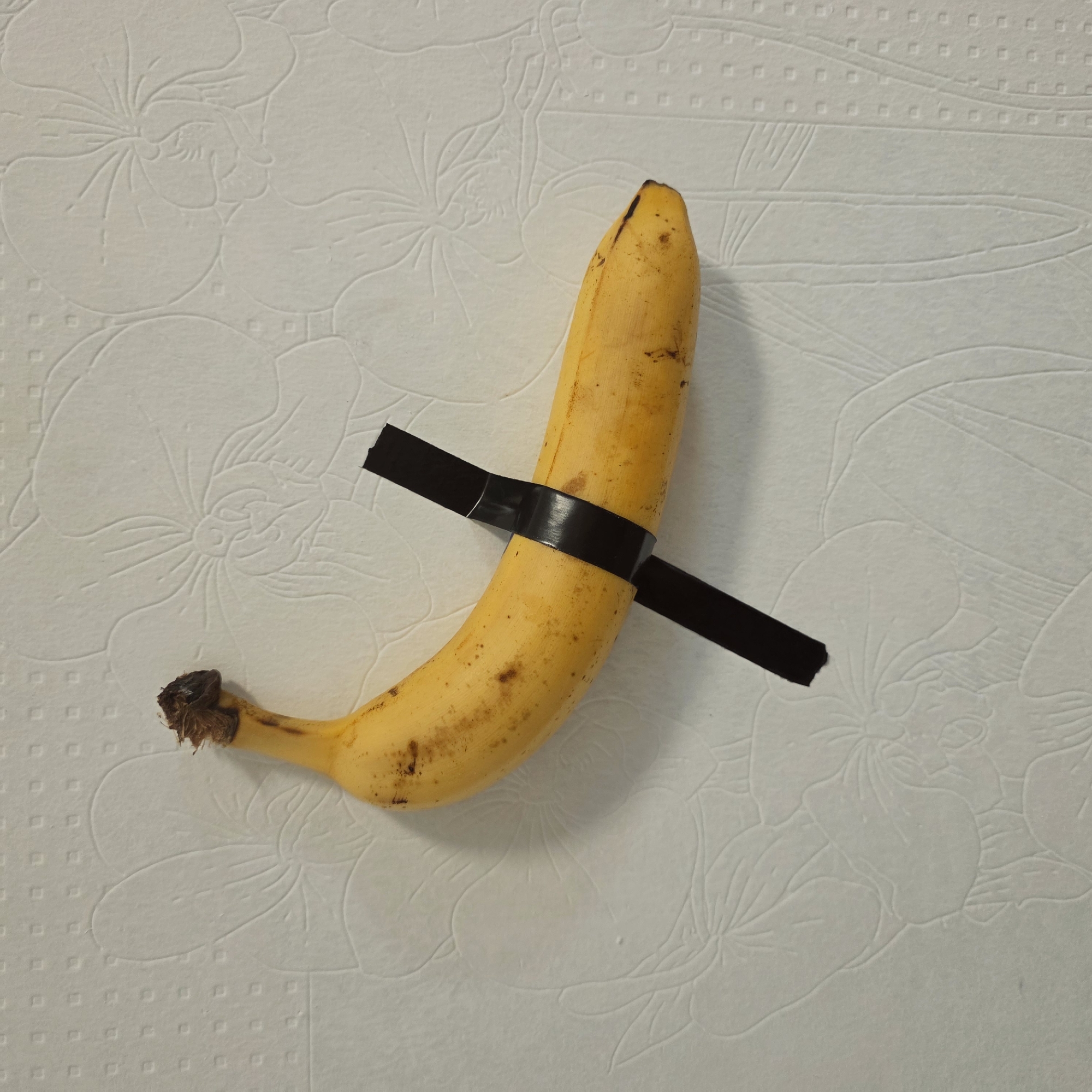A slightly overripe yellow banana with brown spots is taped to a textured, off-white wall with a strip of black duct tape. The banana is positioned vertically on the wall and secured in the middle with the tape that extends from the banana to the wall. The wall appears to be patterned with a raised floral design consisting of flowers. The location is not determined by this image.