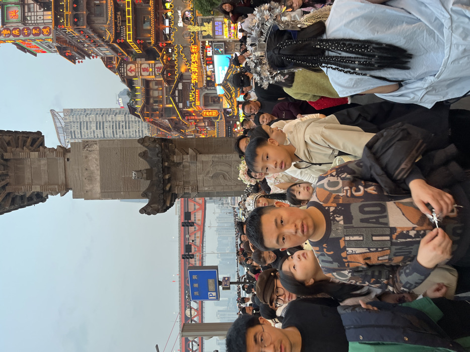 This image depicts a densely crowded street scene at dusk in the Hongyadong Commercial Street, Yuzhong District, Chongqing, China. In the foreground, numerous individuals of East Asian ethnicity fill the frame. A man with short black hair, wearing a multi-patterned long-sleeve shirt in black, orange, beige, and grey, stands centrally, holding a small reflective object in his hands, looking directly ahead. To his immediate left, a man wearing glasses and a black shirt is partially visible. To his right, a young boy in a light-colored hooded sweatshirt looks towards the left. Further right and with her back to the camera, a person wears a light blue garment, elaborate silver headwear, and multiple long, braided hair extensions with silver accents. In the background, to the left, the distinctive red arch of the Hongyadong Bridge spans across water, with distant city skyscrapers visible under a hazy sky. A blue road sign with Chinese characters "洪崖洞" and a 'P' for parking is also visible. To the right, a multi-tiered complex of traditional Chinese-style buildings, characteristic of Hongyadong, is brightly illuminated with numerous golden and red lights and neon signs, creating a vibrant, bustling atmosphere. A tall, ornate grey stone pillar, adorned with carvings, stands prominently in the mid-ground, separating the bridge view from the illuminated buildings. The overall environment suggests a popular tourist or commercial area with heavy pedestrian traffic.