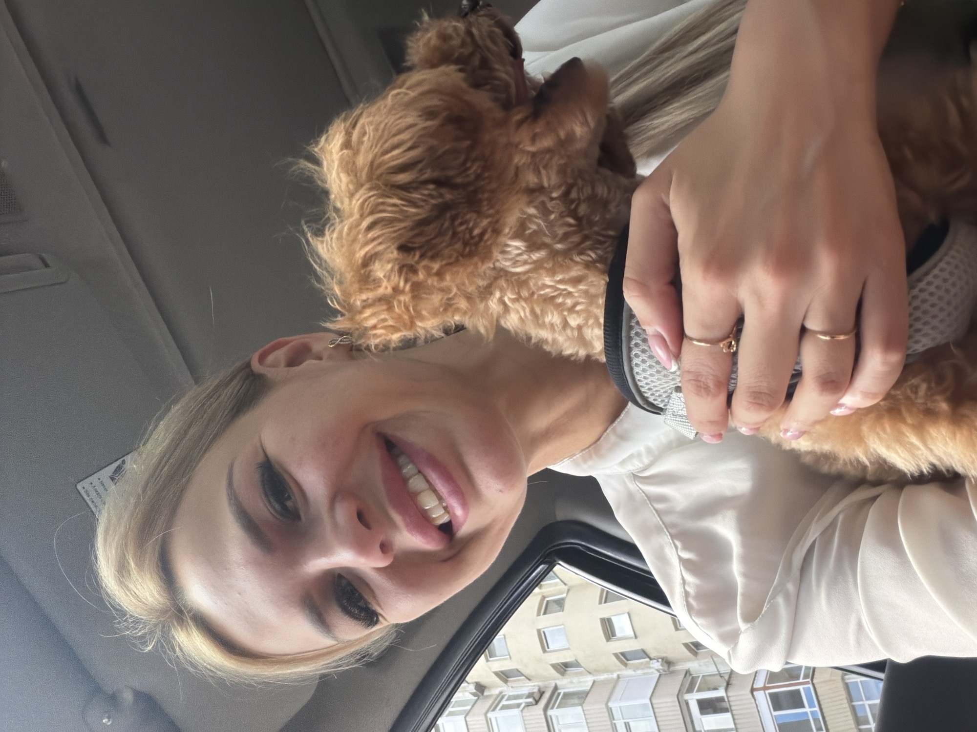 In Kyiv, Ukraine, a woman with blonde hair and a white blouse smiles at the camera while holding a small, brown, curly-haired dog. The dog is wearing a gray harness with white trim. The woman is wearing two gold rings on her left hand, one with a small floral design. In the background, through the car window, a multi-story residential building with light-colored walls and numerous windows is visible. The sky appears overcast.