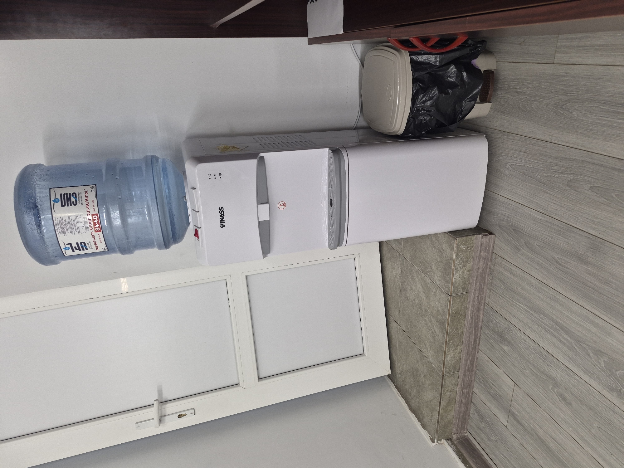 A white water cooler stands next to a white door and a small trash can. The water cooler has a blue water bottle on top and a dispenser at the bottom. The trash can is beige with a black trash bag inside. The door is closed. The floor is made of grey wood-like planks. The scene is indoors, likely in an office or public space. There is no indication of the location being specifically in Yerevan, Armenia.