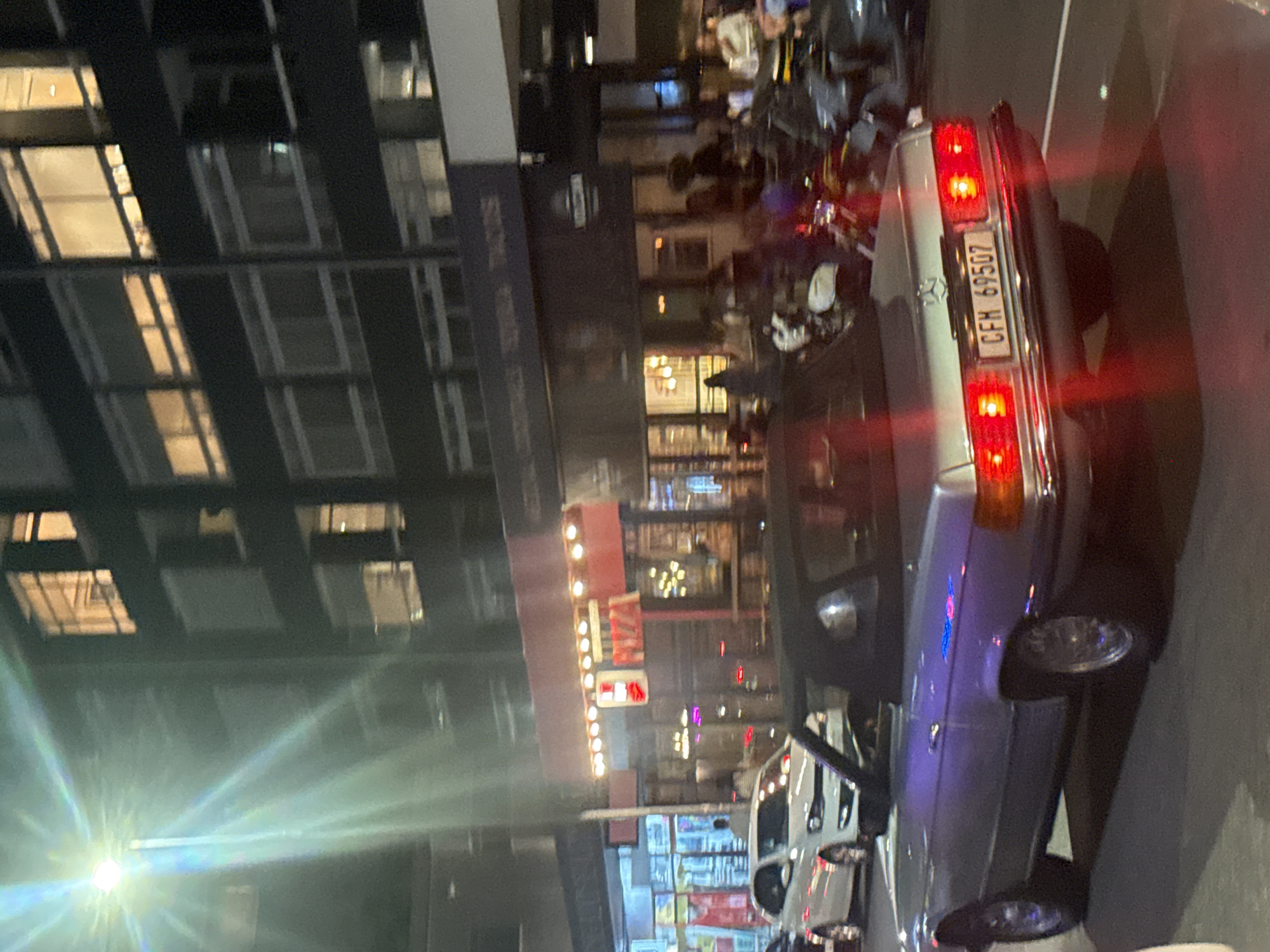 The image depicts a street scene at night in what appears to be the city of Cape Town, South Africa. The focus is on a silver-colored vintage Mercedes-Benz parked along the curb. The car's license plate is visible, reading "CFM 69507". The rear lights of the car are illuminated. A row of restaurants with outdoor seating areas is visible behind the car, suggesting a lively atmosphere. People are gathered at tables and walking in the area. Modern office buildings are visible in the background, illuminated by interior lights. A street light is shining brightly, creating a starburst effect. The street is wet and reflective, suggesting recent rain. Several other cars are parked in the vicinity.