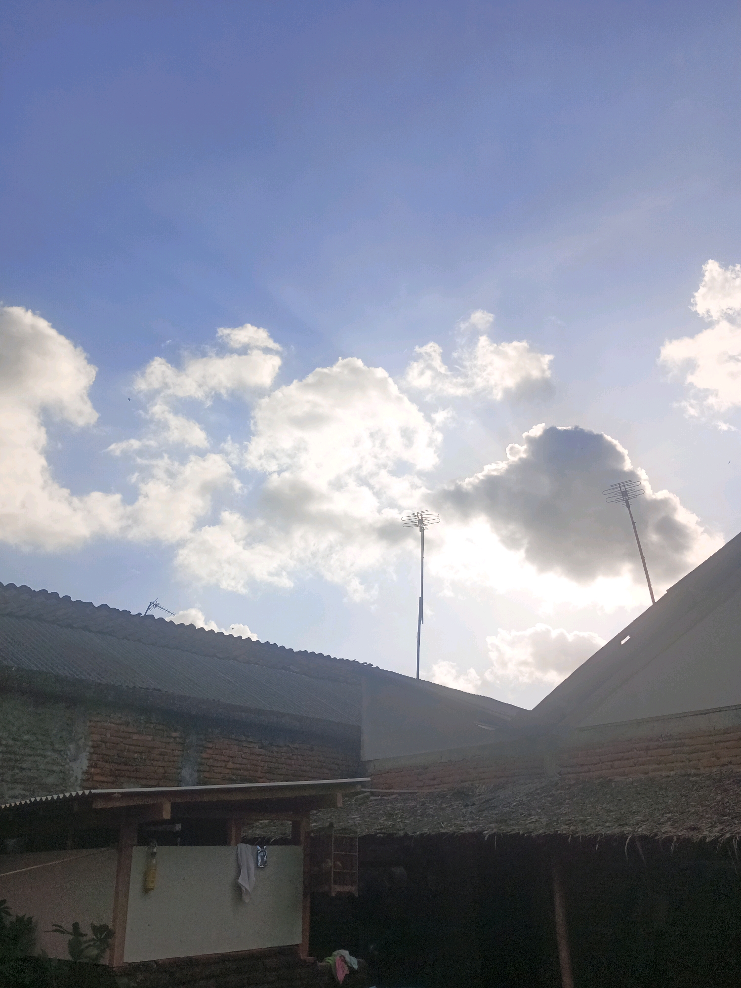 The image captures a cloudy sky with patches of sunlight breaking through, illuminating two television antennas rising above the rooftops of several buildings. The architecture appears to be of a residential nature, with corrugated metal and tiled roofs visible. One building on the left has a visible brick wall. The scene suggests an outdoor setting, likely in a residential area of Kawunganten, Indonesia. The lighting indicates it may be early morning or late afternoon. There are no people visible in the frame.