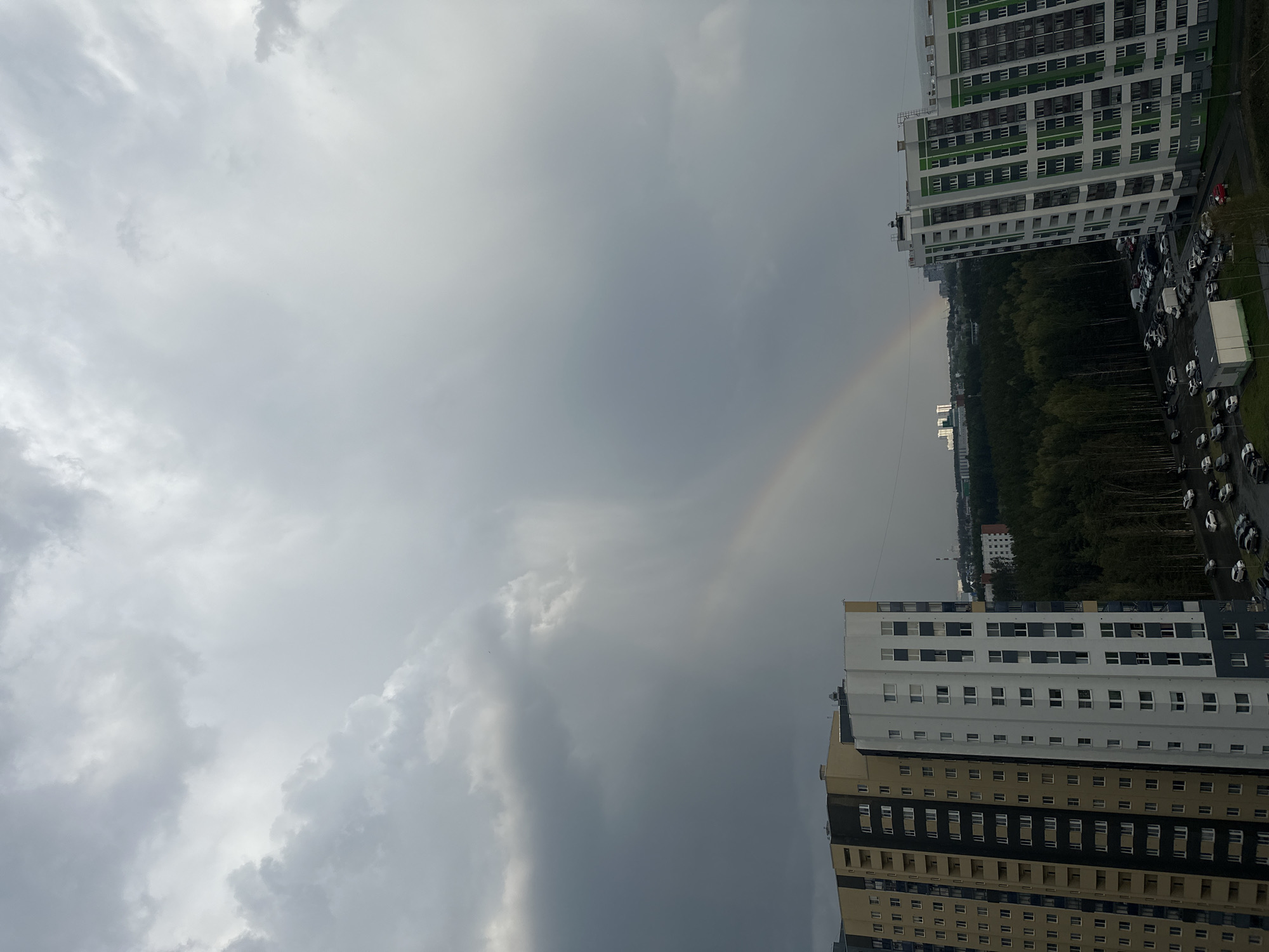 The image shows a cityscape under a cloudy sky with a faint rainbow. Two tall apartment buildings flank the central view. One building is predominantly white with green accents, while the other is a mix of beige, grey, and black. Between them, a line of trees and parked cars is visible, leading toward smaller buildings in the distance. The heavy cloud cover dominates the sky, creating a somber atmosphere. This scene is located in the city of Yekaterinburg, Russia.