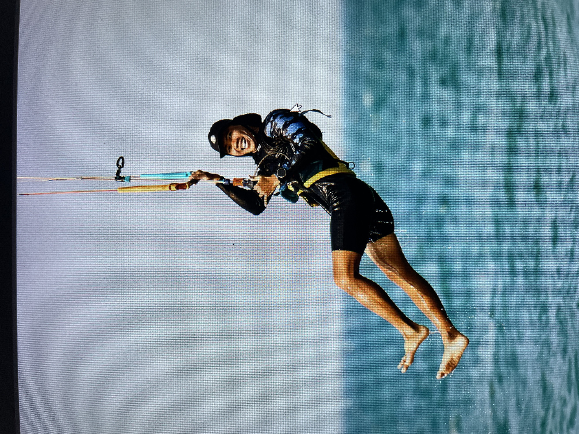 A woman is kitesurfing. She is airborne, holding a kite control bar. The background shows light blue sky on the left and slightly choppy water on the right.
I am unable to locate the city of Bacong in the image and cannot identify it as the location.