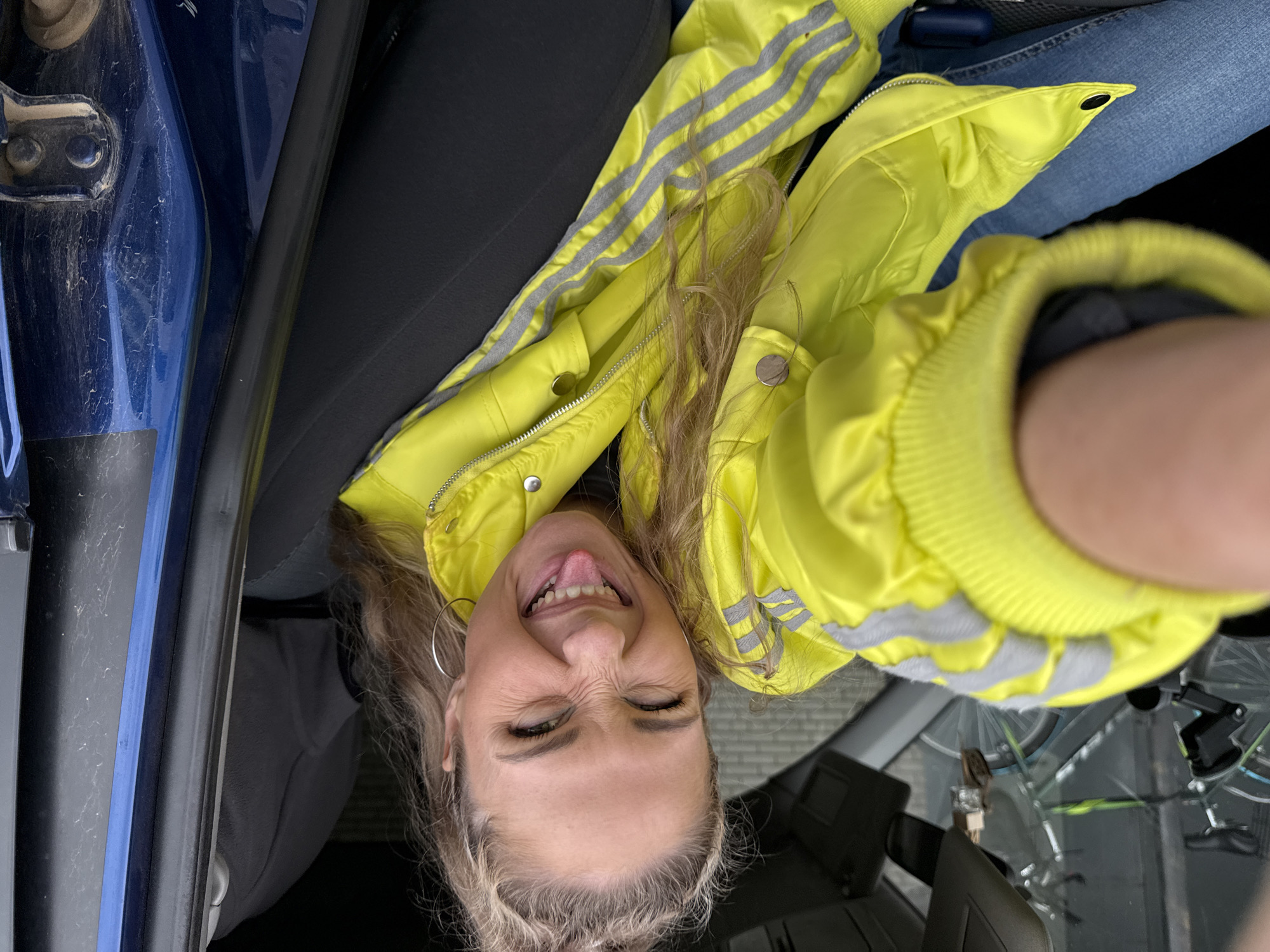 A woman with blonde hair pulled into a high ponytail is sitting in the passenger seat of a vehicle, wearing a bright yellow jacket with gray stripes on the sleeves and blue jeans. She is making a funny face, sticking out her tongue and squinting her eyes. The interior of the vehicle is visible, including the car seat and the car door. Outside the vehicle, a bicycle is visible through the windshield. The location is identified as Chaikovsky, Russia.