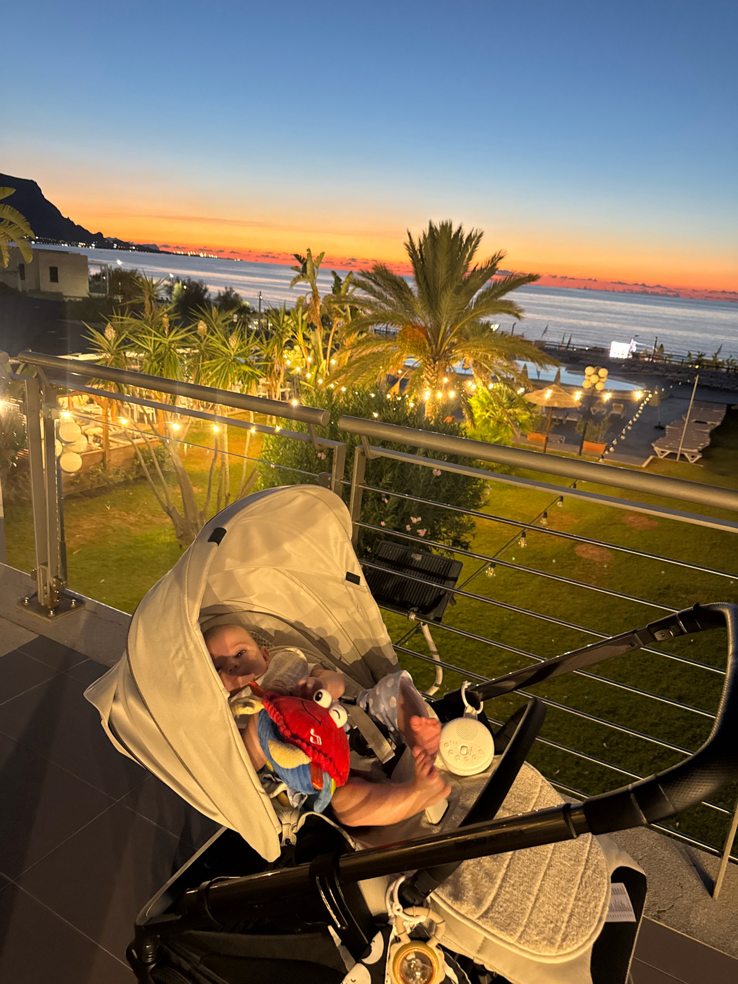 A baby is in a stroller on a balcony overlooking a body of water and a coastline with lights. The sky is a gradient of blue, orange, and pink, indicating either sunrise or sunset. Palm trees and other greenery are visible, along with outdoor lights strung across the balcony and in the distance. The stroller has a canopy and a plush toy. The location is Isola delle Femmine, Italy.