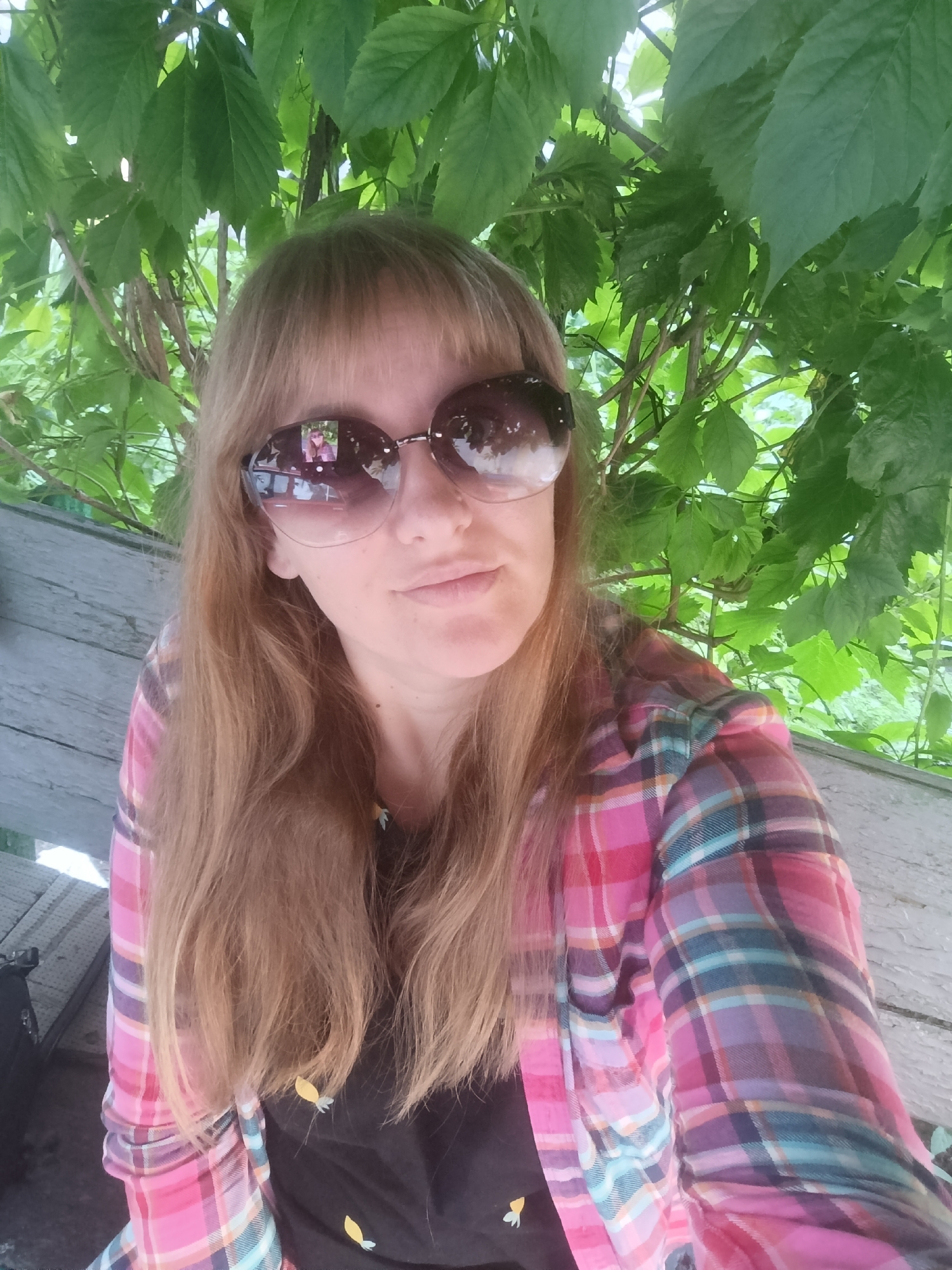 A woman with long, blonde hair and bangs, wearing large sunglasses, a pink and blue plaid shirt, and a black t-shirt with yellow flower designs, is pictured from the chest up. She is sitting on a weathered wooden bench outdoors, with green foliage overhead and to the right. The reflection in her sunglasses shows herself and what appears to be a railing. The bench is to the left of the frame, and a dark object, possibly a bag, is visible at the bottom left corner.