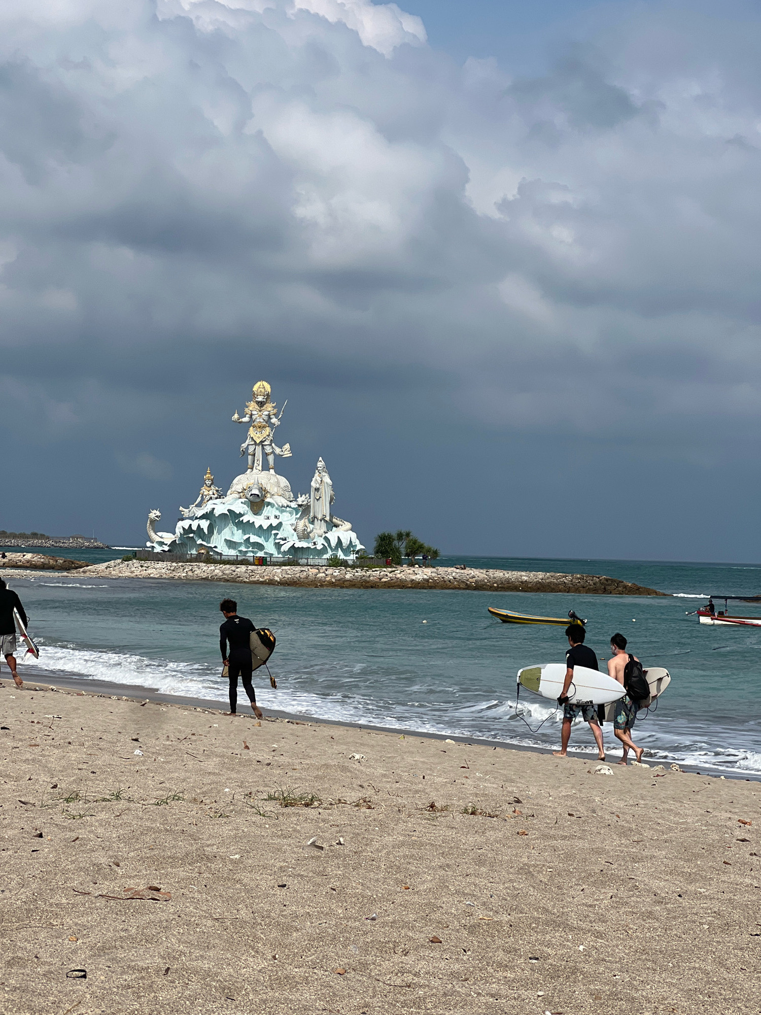 The image depicts a beach scene in Kuta, Indonesia, with a large statue of a deity on a rocky island in the background. The statue is adorned in gold and appears to be a religious or cultural monument. The sky is overcast with dark clouds, suggesting a potential for rain.
In the foreground, three individuals are visible on the sandy beach. Two of them are carrying surfboards and are walking along the shoreline, appearing to be surfers. One individual is wearing a full wetsuit, while the other is wearing shorts and a t-shirt. The third person is further up the beach and is also carrying a surfboard.
The ocean is a vibrant blue-green, with gentle waves breaking on the shore. There are a couple of boats visible in the water near the statue and further out to sea. The beach itself is sandy with some scattered debris, including dried leaves and small stones. The overall atmosphere of the image is one of a typical beach day with a prominent cultural landmark in the distance.