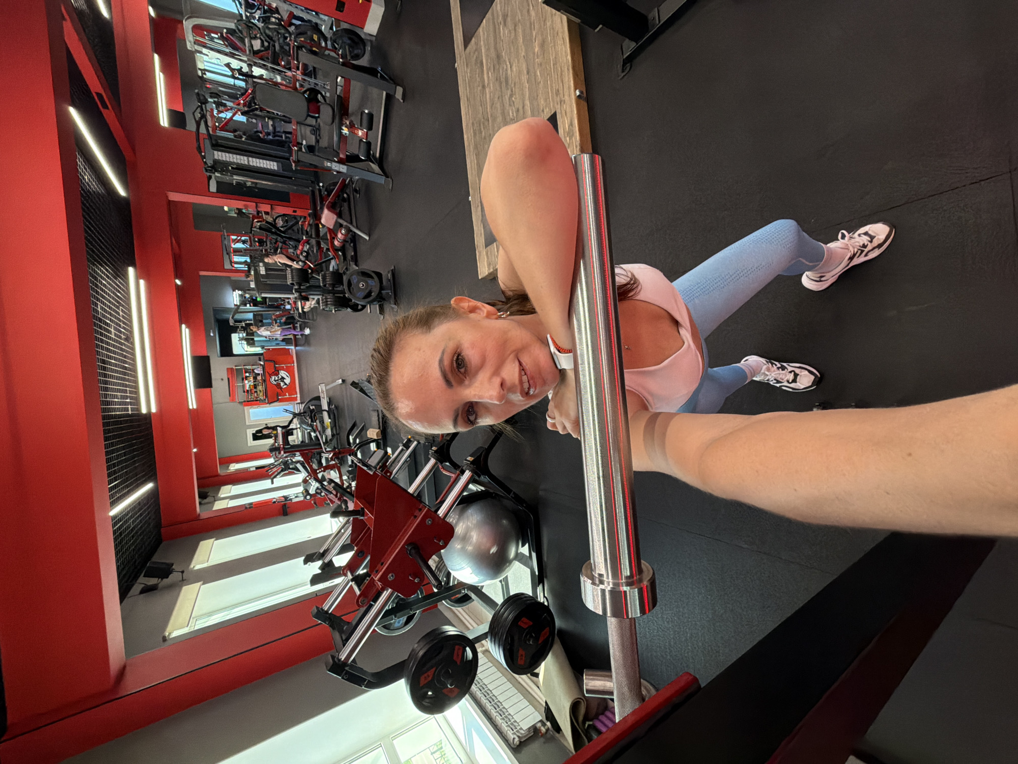 This is a selfie taken by a woman in a gym. The woman is wearing a pink tank top and blue leggings, with white and black athletic shoes. She is holding a barbell, which appears to be part of a squat rack, and is in a squatting position. The gym has red accents on the ceiling and equipment. In the background, there are various weightlifting machines and a large fitness ball. The flooring is black rubber. The lighting is artificial, with fluorescent lights visible on the ceiling. Based on the context provided, this gym is located in Birobidzhan, Russia.