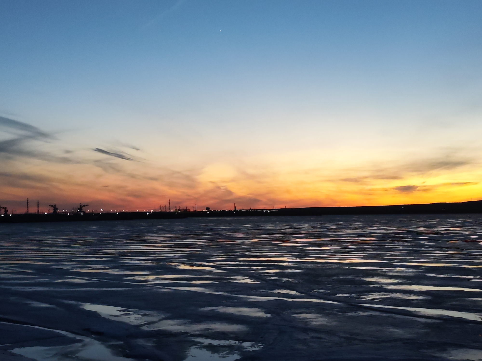The image depicts a landscape at sunset over what appears to be a partially frozen body of water, possibly in or near Chaikovsky, Russia. The sky transitions from a deep blue at the top to shades of orange and yellow near the horizon, with some streaky clouds visible. In the distance, there is a silhouette of what may be industrial structures and towers against the brightly lit sky, possibly indicating a cityscape or industrial area. The foreground is occupied by the frozen water surface, showing dark and light patterns that suggest a mix of ice and water reflecting the colors of the sky.