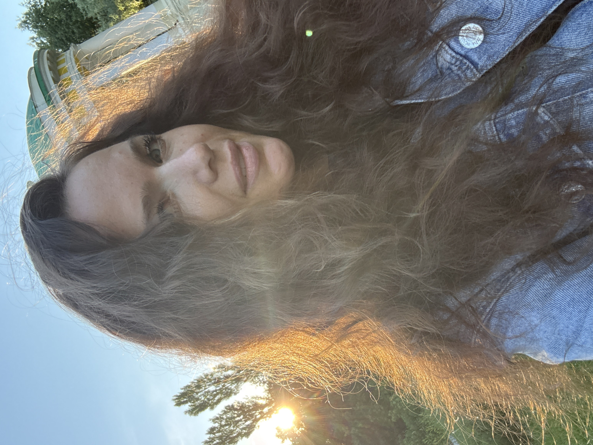 The image shows a close-up, low-angle selfie of a young woman with long, dark brown hair. She is wearing a blue denim jacket. Her face is partially obscured by her hair, but her left eye, nose, and mouth are visible. She appears to be looking directly at the camera with a neutral expression. The setting appears to be outdoors, with a bright sun low in the sky, casting a warm golden light on her hair. The sky is a clear blue, and some green foliage is visible in the background. The overall mood of the image is casual and relaxed. It is difficult to discern any specific text in the image, other than a small emblem on the button of her jacket. The time of day appears to be either early morning or late afternoon due to the low angle of the sun. The location context is Moscow, Russia.