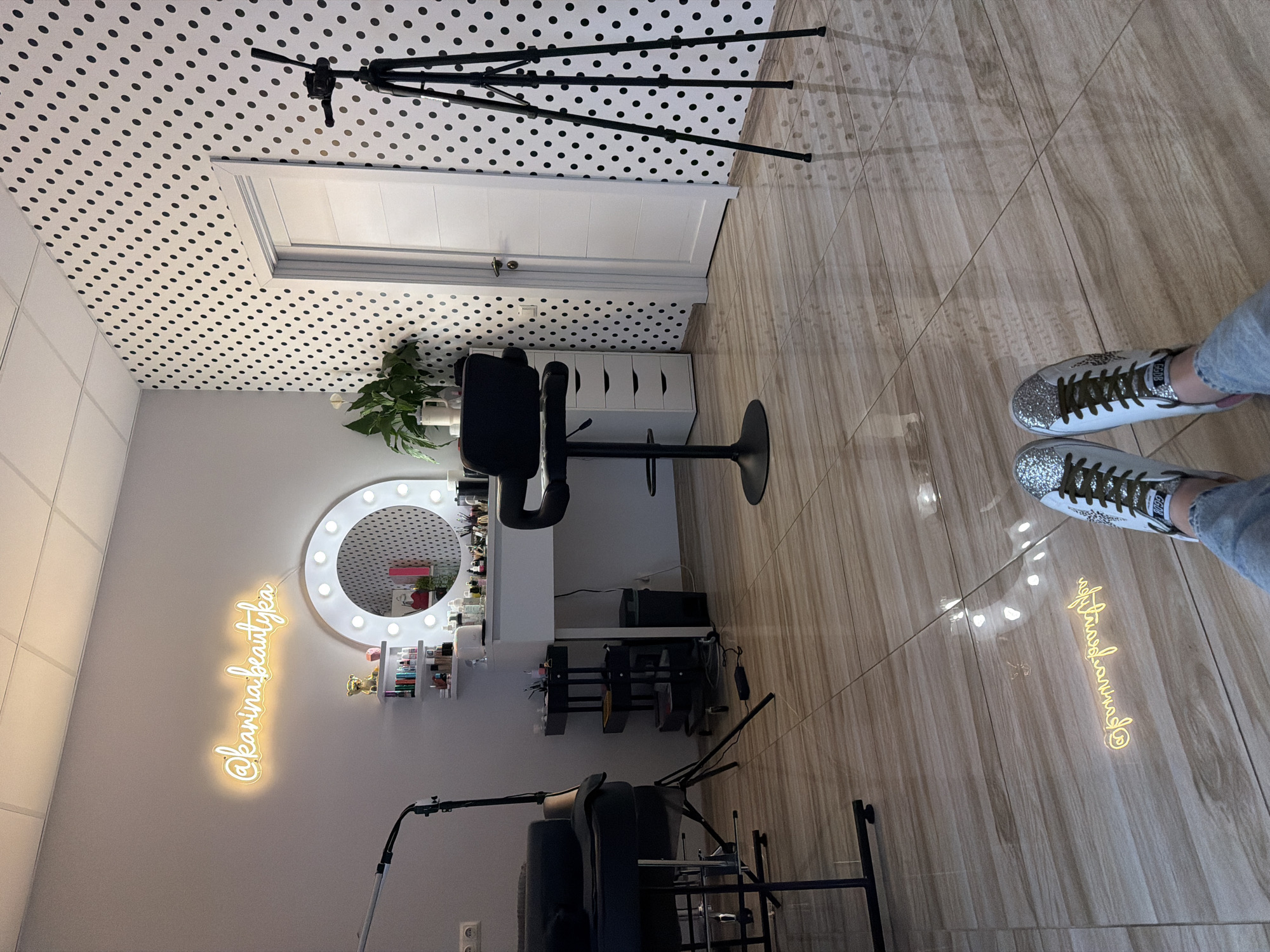A photograph depicts an interior space, likely a professional makeup or beauty studio. The space is observed from an elevated perspective. The walls are light gray, with one wall exhibiting a black and white polka dot wallpaper pattern that extends onto the ceiling. A white door is situated within the polka-dotted wall.
The room features a makeup vanity with a circular, illuminated mirror. Various makeup products and decorative items are arranged on shelves surrounding the mirror. A black adjustable stool is positioned in front of the vanity. Additional storage in the form of white drawers is situated to the right of the makeup area.
Several pieces of equipment, including tripods and lighting rigs, are present. The floor is tiled with a light brown, wood-like pattern.
The lower right corner of the image shows a pair of feet clad in gray jeans and white sneakers with green laces and glittery embellishments. On the floor is a mirrored projection of a neon sign that reads "@karinabeautykea" in cursive script.
The image was taken in Riga, Latvia.