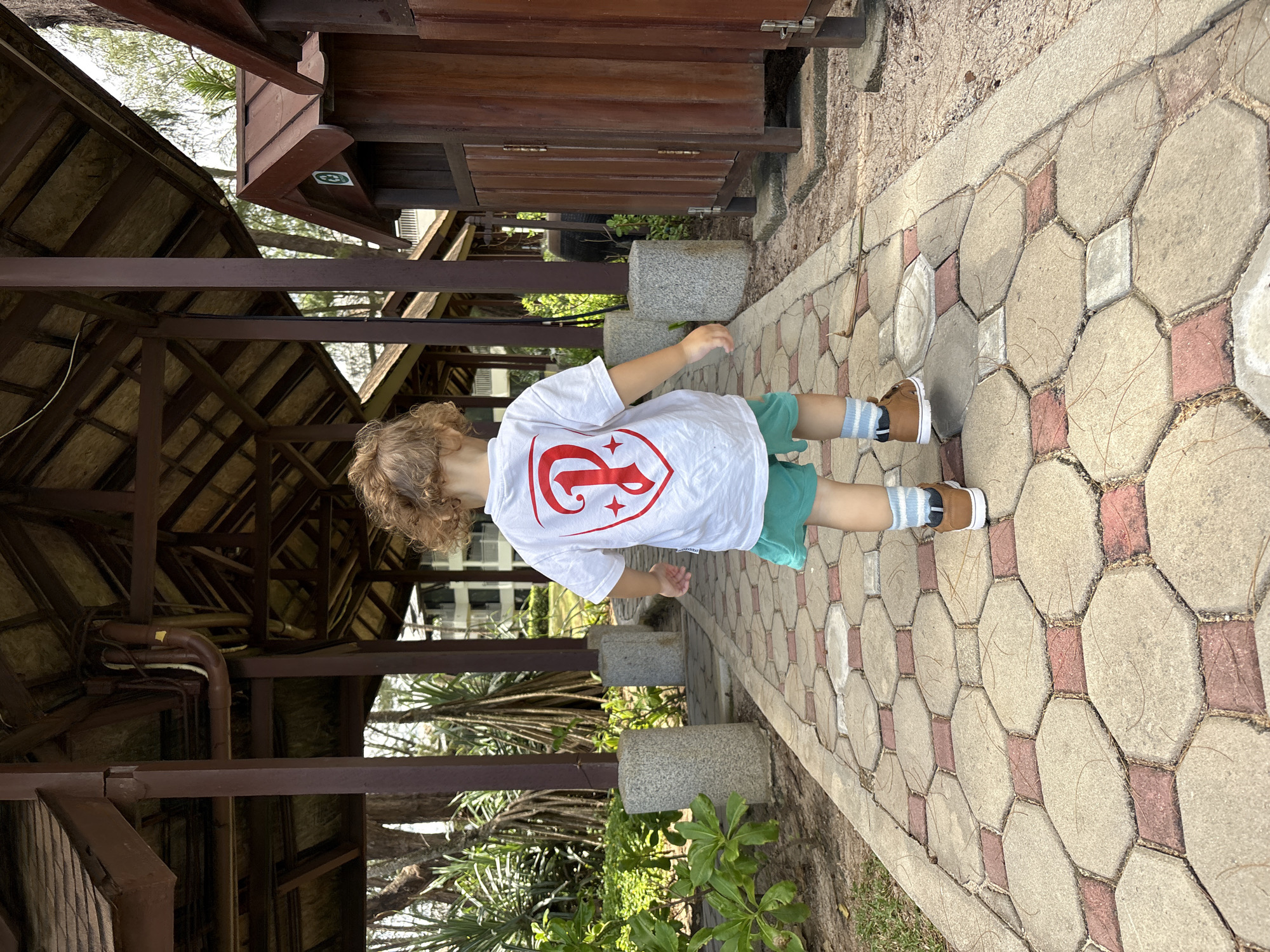 A child with curly brown hair is walking away from the camera on a paved pathway in Choeng Thale, Thailand. The child is wearing a white t-shirt with a red emblem on the back, green shorts, blue and white striped socks, and brown shoes. The pathway is made of interlocking octagonal and square concrete pavers with reddish-brown grout. The path is lined with greenery and shaded by a wooden structure with a thatched roof, supported by square wooden posts. To the right of the pathway are wooden structures that appear to be shower stalls or changing rooms. In the background, trees and a building can be seen. The ground on the sides of the path is sandy and covered with pine needles.