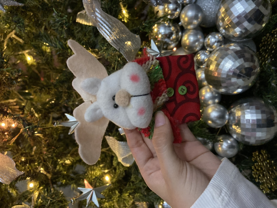A person holds a handmade felt Christmas ornament shaped like a white winged reindeer with rosy cheeks and a button nose. The ornament is attached to a small red pouch adorned with two green buttons. The backdrop features a decorated Christmas tree with a mix of silver ornaments, including disco balls and ribbon. The lighting is soft, creating a warm festive atmosphere.